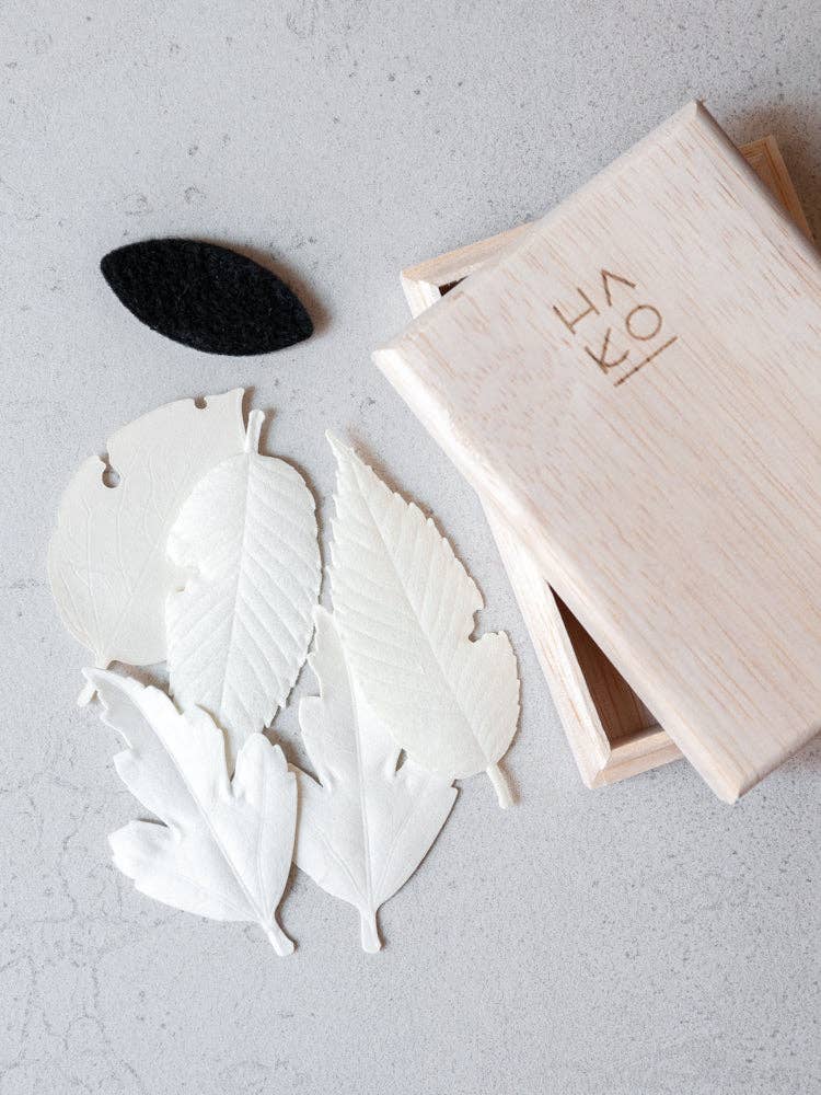 A wooden box with Japanese characters contains HA KO Paper Incense—several white paper leaves and one black felt leaf—displayed on a light gray surface, making an elegant Japanese incense gift set.