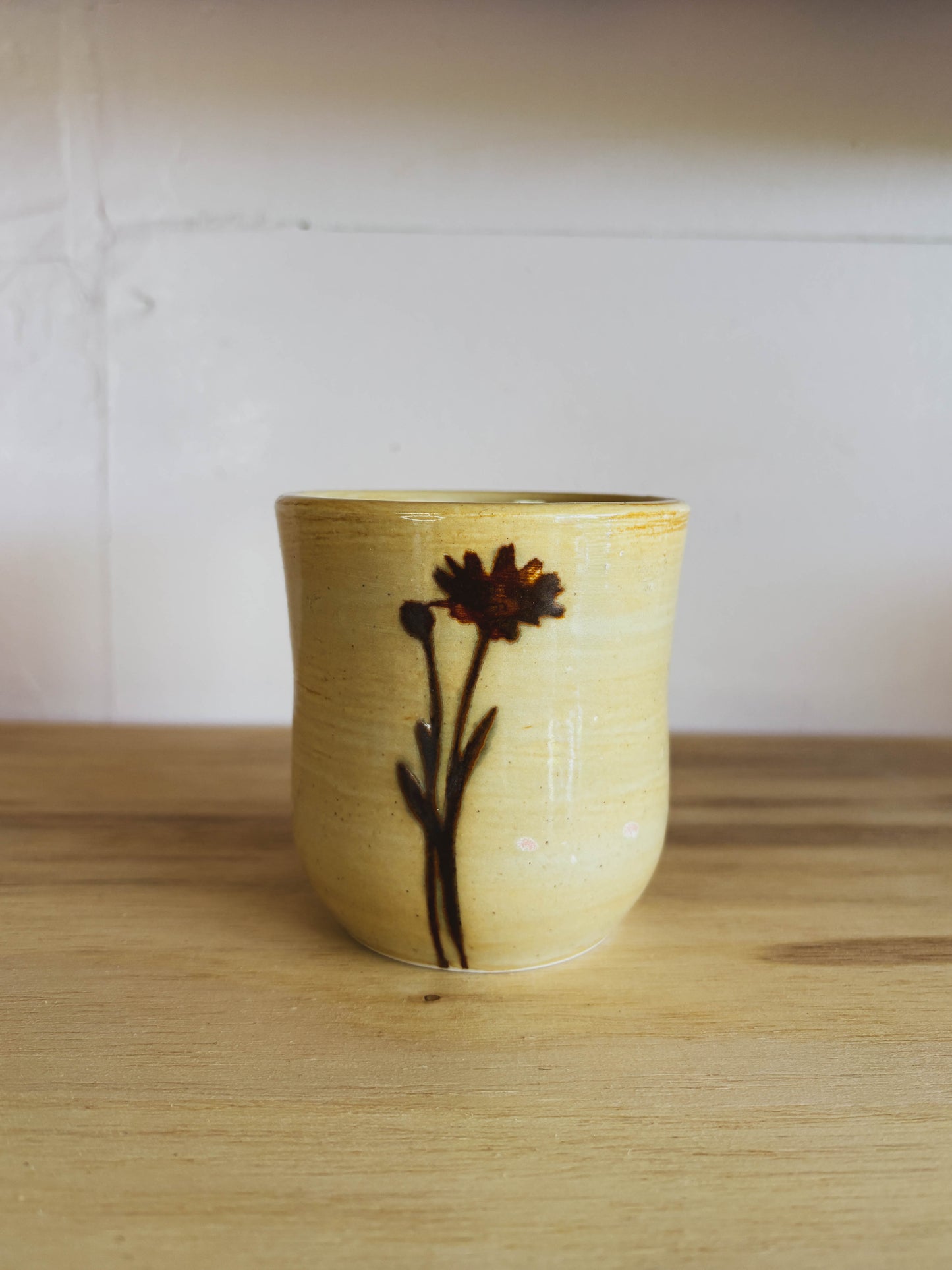 The Birch Arnica #1 Small Tea Cup is a beige, handcrafted ceramic cup featuring a simple brown flower design, shown on a wooden surface with a white wall in the background.