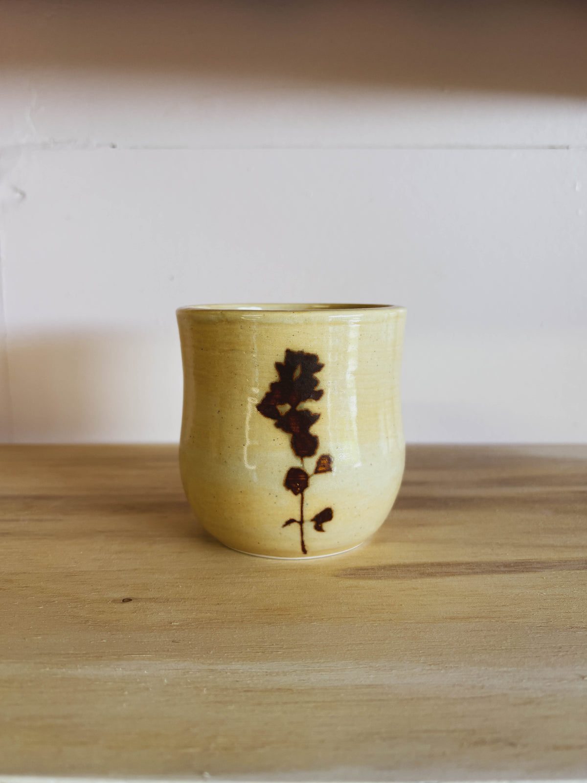 The Birch Globemallow #1 Medium Tea Cup is a handmade yellow ceramic cup featuring a simple brown floral design, displayed on a wooden surface with a pale pink wall in the background.
