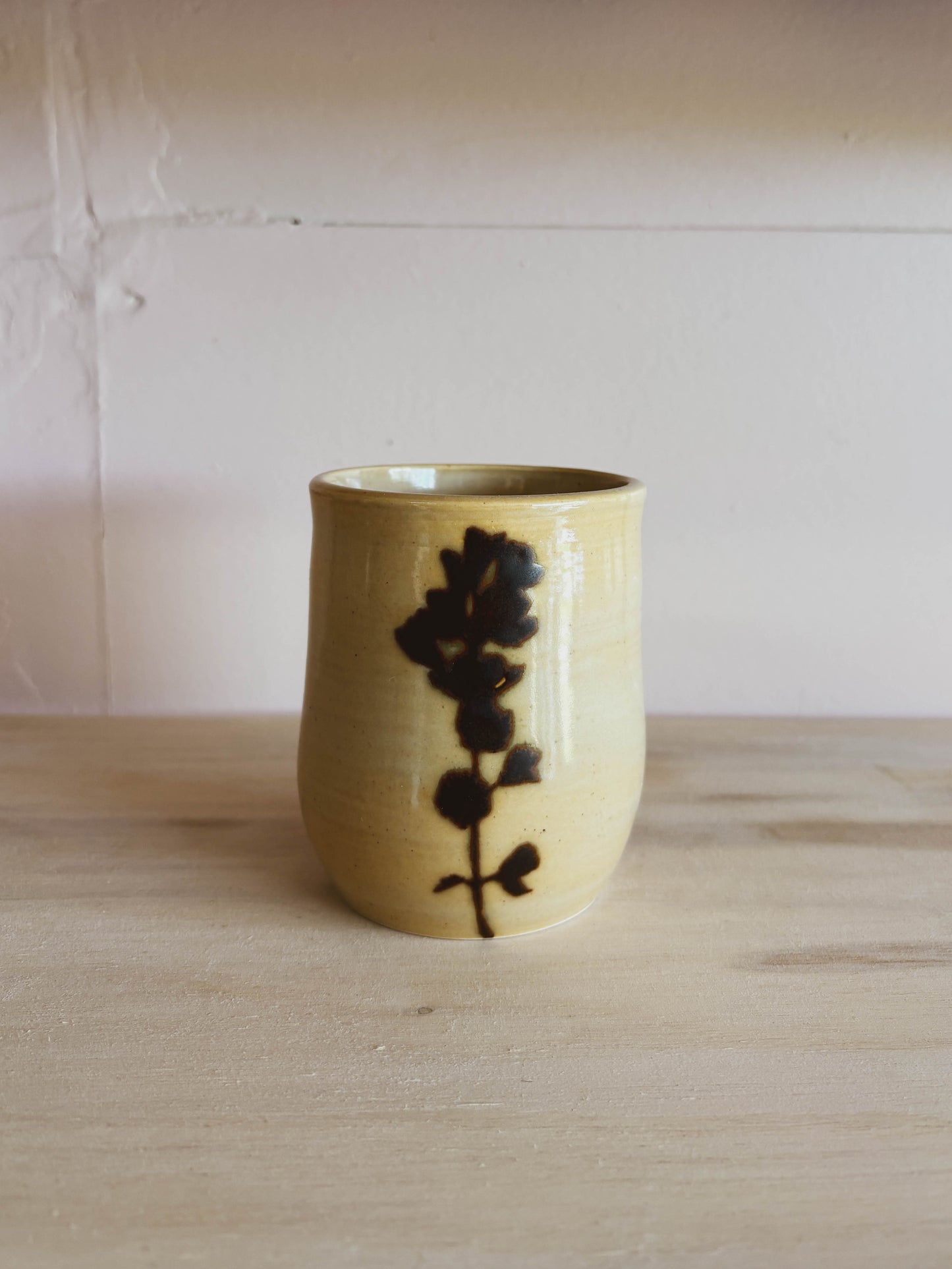 The Birch Globemallow #2 Medium Tea Cup, handcrafted from beige ceramic with a hand-painted dark floral design, rests on a light wooden surface against a pale, textured wall.