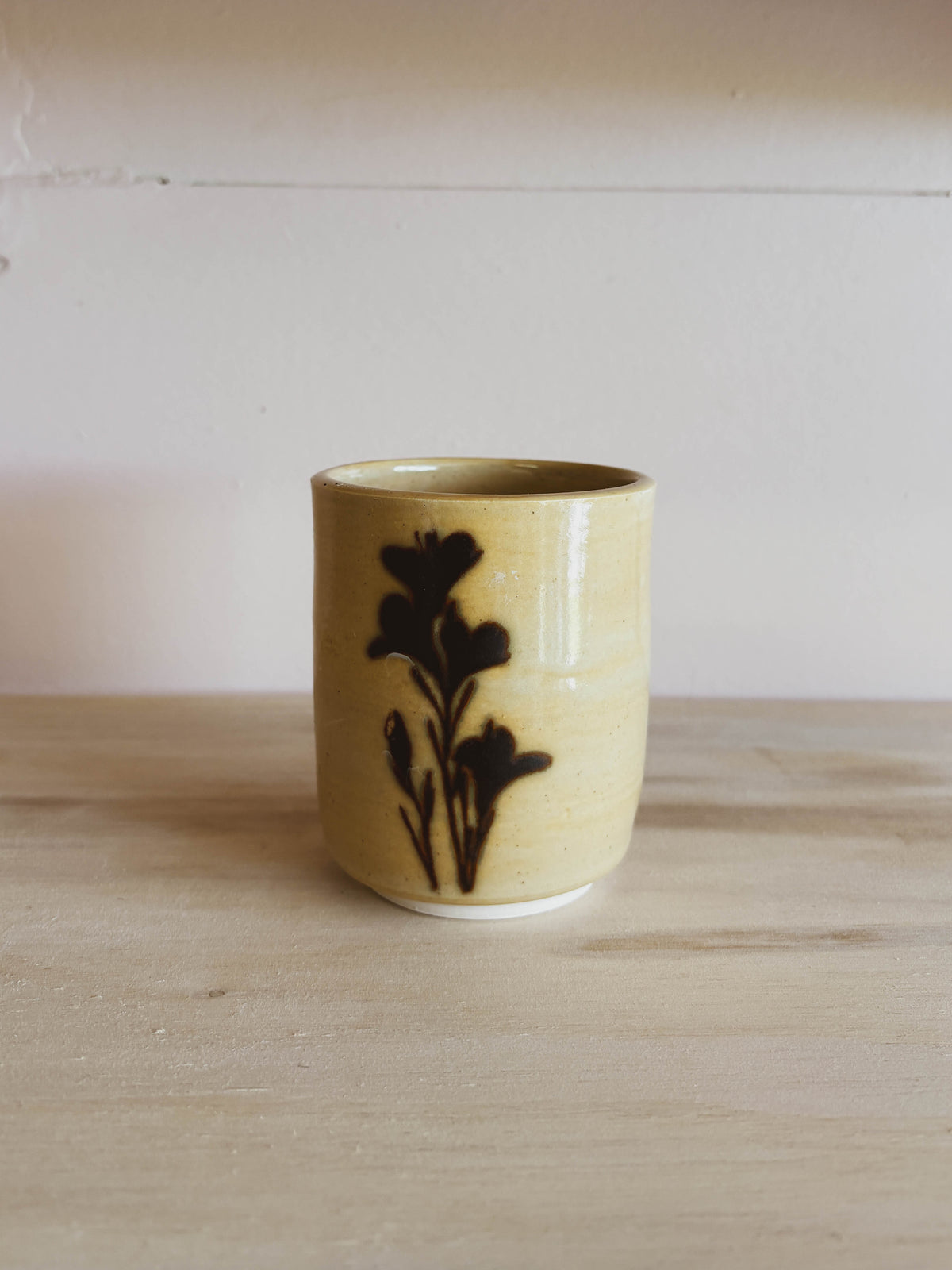The Birch Lily #1 Small Tea Cup, featuring a handcrafted ceramic build with a dark brown floral design, rests on a light wooden surface against an off-white background.