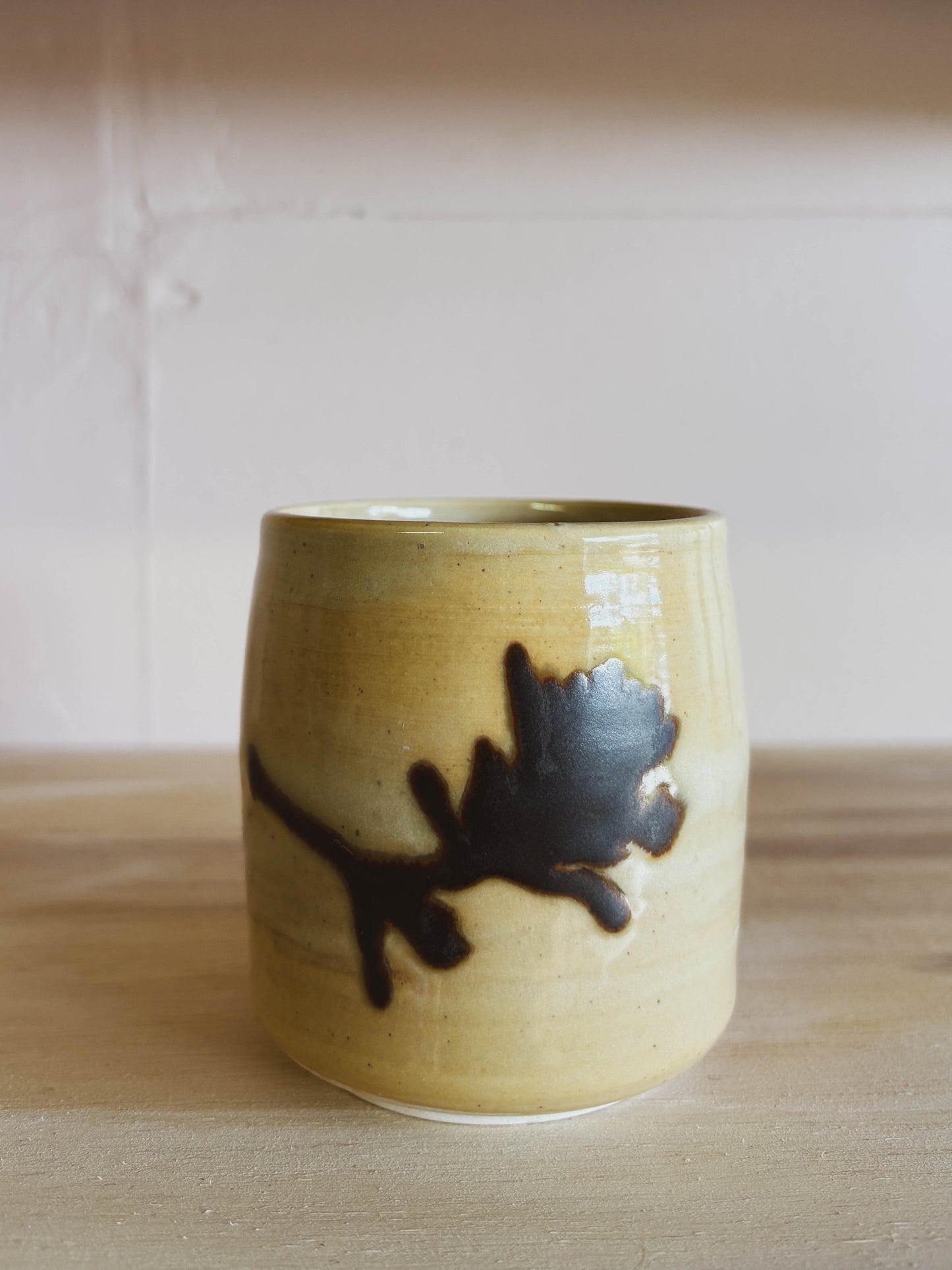 The Birch Paintbrush #1 Small Tea Cup is a handcrafted ceramic cup with a glossy beige finish, dark brown branch and leaf design, dishwasher safe, made by a women-owned business. It’s shown on light wood against a pale background.