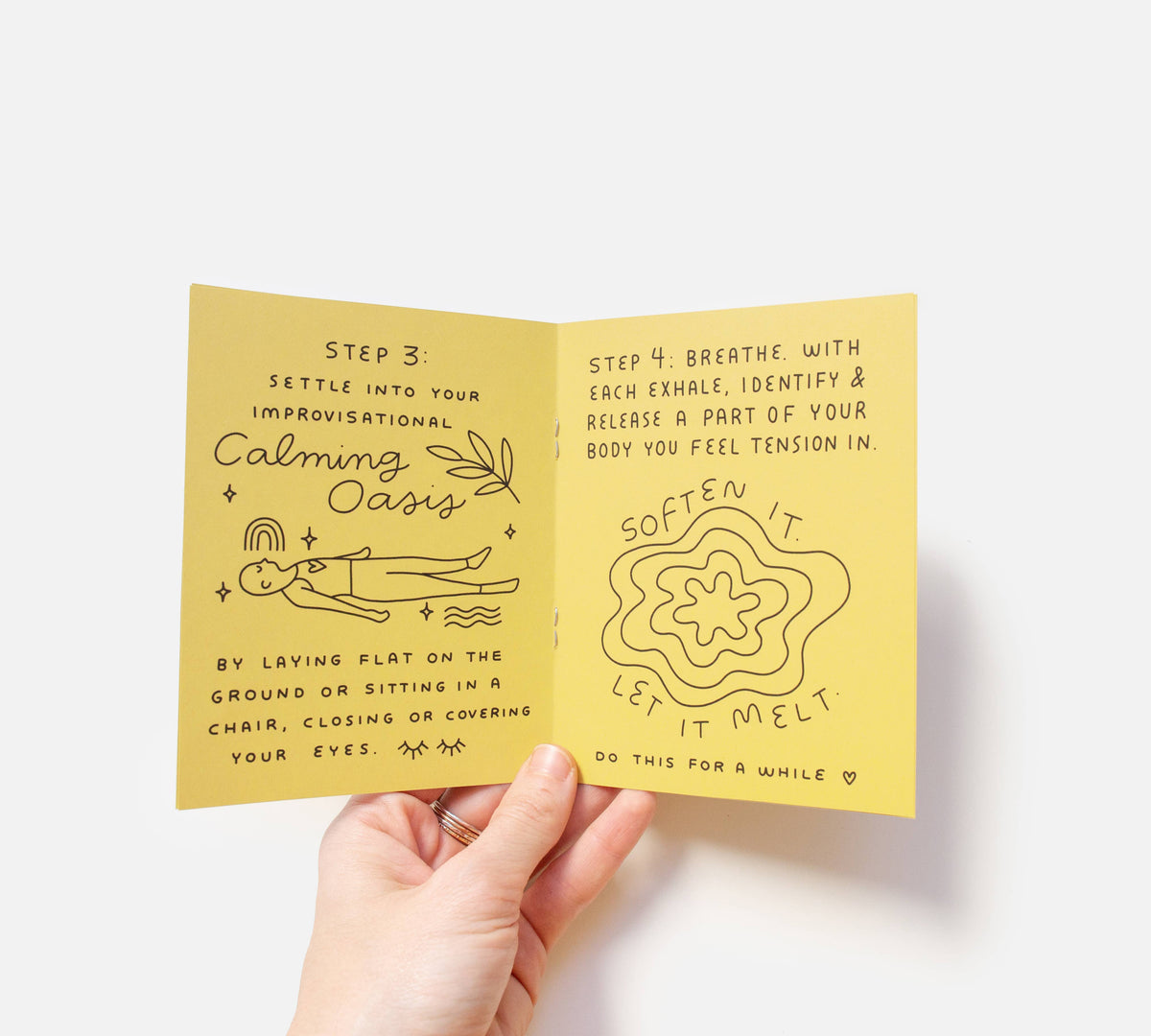 A hand holds an open "Take a Break Zine," a yellow handmade booklet with calming illustrations and relaxation instructions, such as breathing tips and releasing body tension, featuring soothing text and peaceful imagery.