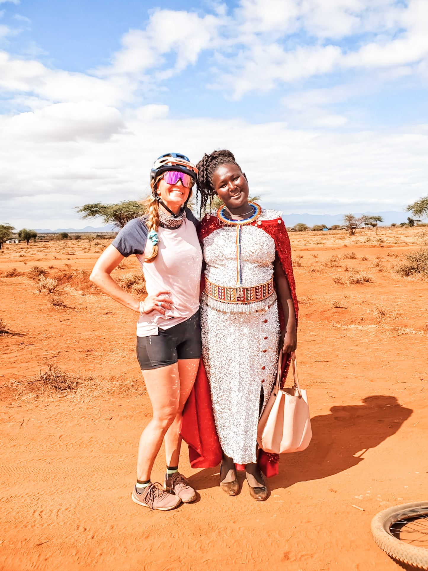 Two women pose together on a sunny, red-dirt landscape with scattered trees: one wears Cycling Safari gear and a helmet, while the other is in traditional Maasai attire with beaded jewelry and a red cape.