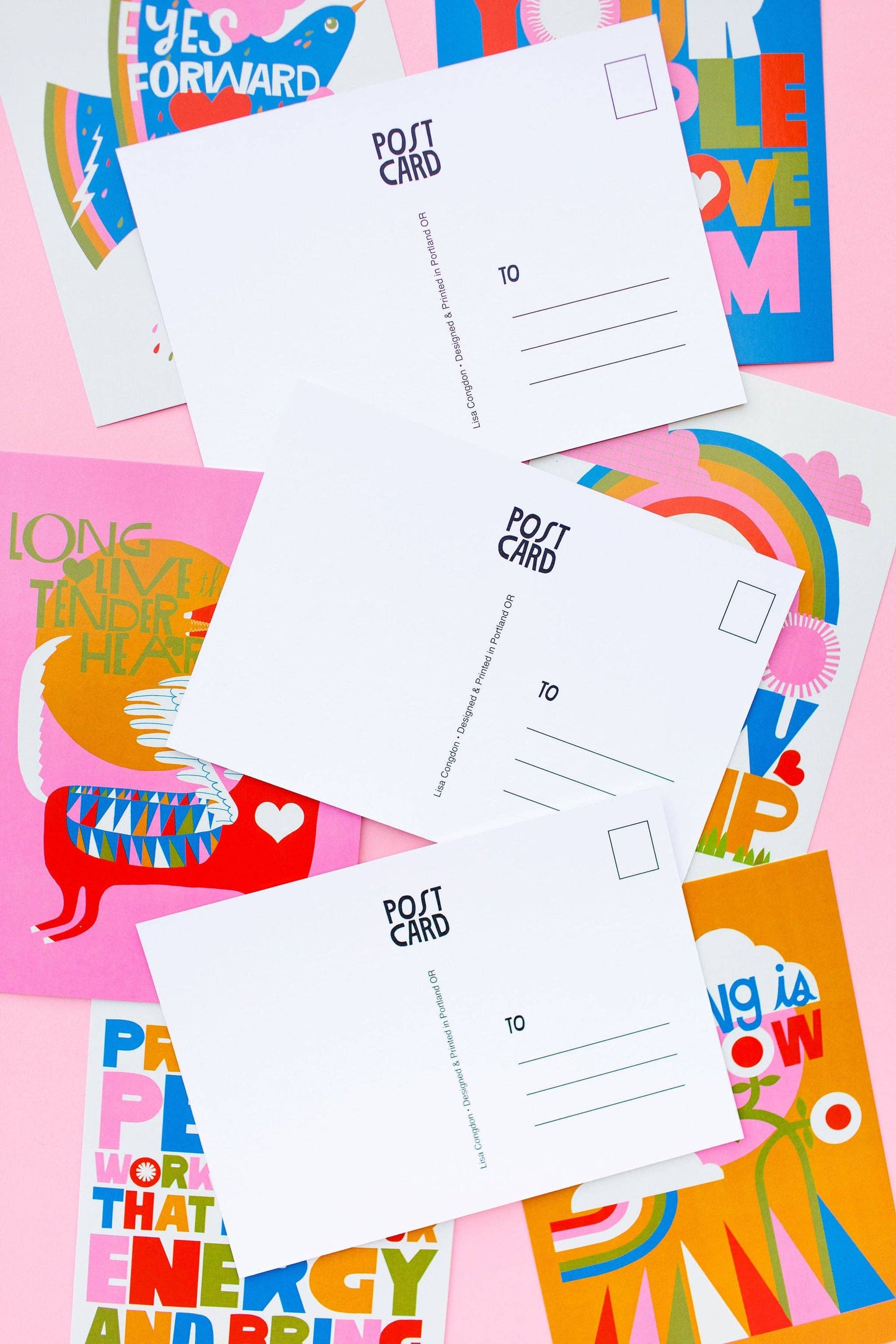 Three blank postcards from the XL Postcard Set are displayed atop colorful, illustrated greeting cards with encouraging messages and bold designs in pink, blue, orange, and yellow—each uniquely printed in Portland.