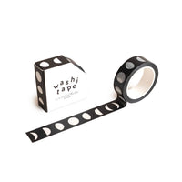 The Moons Washi Tape, an eco-friendly black tape featuring a striking white lunar phases pattern, is partially unrolled beside its matching "washi tape" box for a stylish and sustainable display.