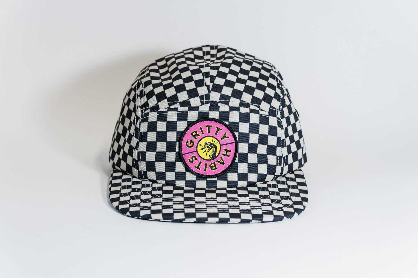 The Steezy Checkered 5-Panel Hat features a black and white checkered design with a pink "GRITTY HABITS" patch around a yellow smiley face, made by a women-owned small business. Shown on a crisp white background.