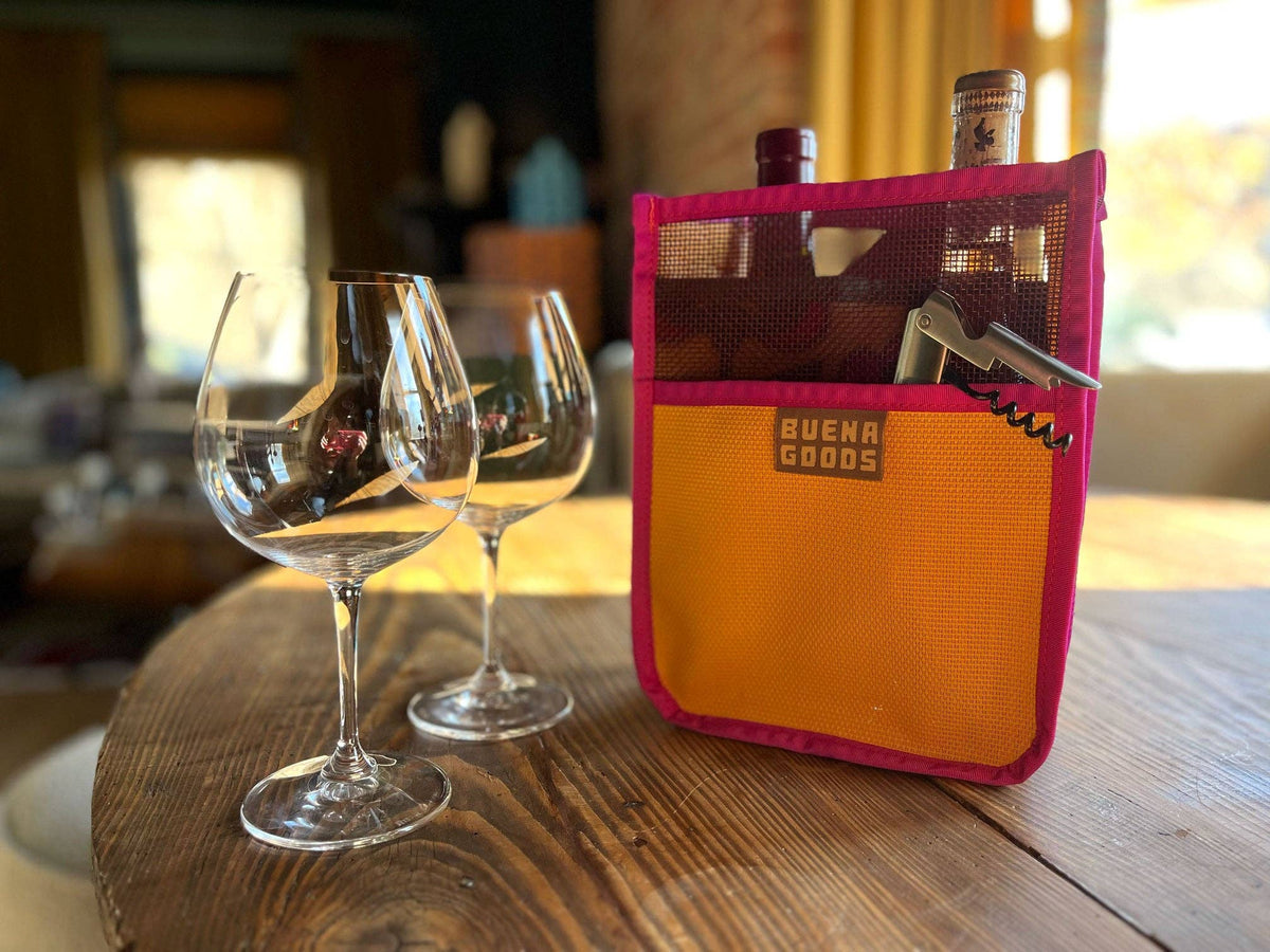 Two empty wine glasses rest on a wooden table beside The Two-Bottle-Bag | Cava on the Playa from Buena Goods, holding wine bottles and a corkscrew, in a cozy, sunlit room.