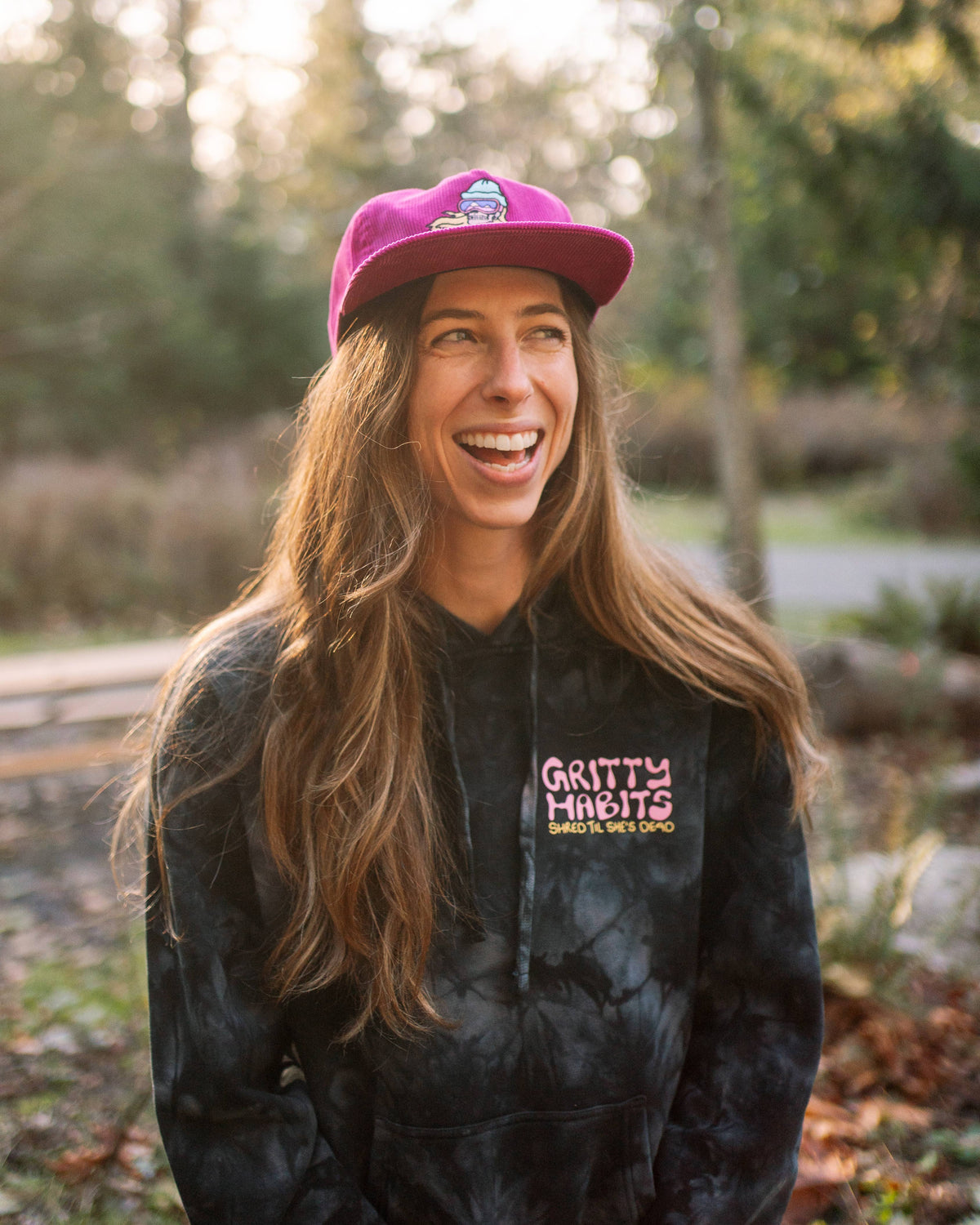 A smiling person with long brown hair wears the Shred Til She's Dead Corduroy 5-Panel Hat and a black "Gritty Habits" tie-dye hoodie, representing a women-owned small business. They stand outdoors among trees and sunlight.