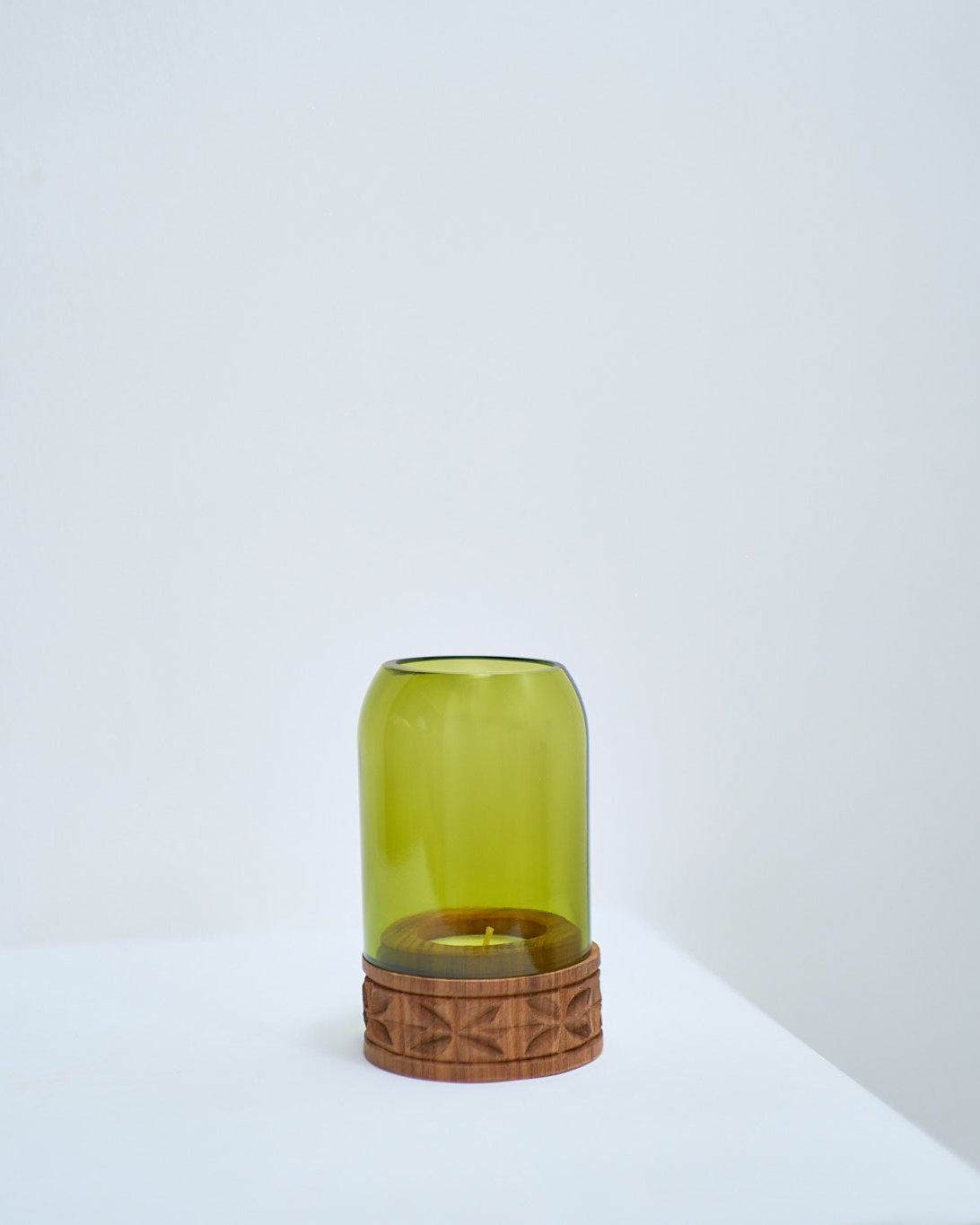 The Upepo Tea Light Holder, made from upcycled green glass with a carved wooden base, rests on a white surface against a plain light-colored wall.