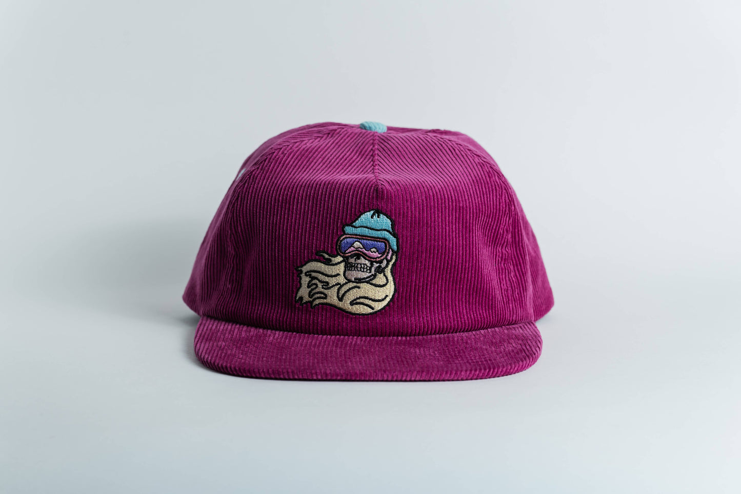 The Shred Til She's Dead Corduroy 5-Panel Hat features magenta corduroy, a patch with a cartoon skull in ski goggles and a blue beanie, wavy line detail, and moisture-wicking sweatband—made by a women-owned small business.