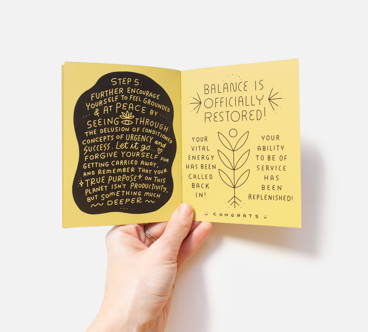A hand holds open the eco-friendly, handmade Take a Break Zine. The left page features grounding and self-forgiveness tips, while the right shows line art declaring that balance is officially restored.