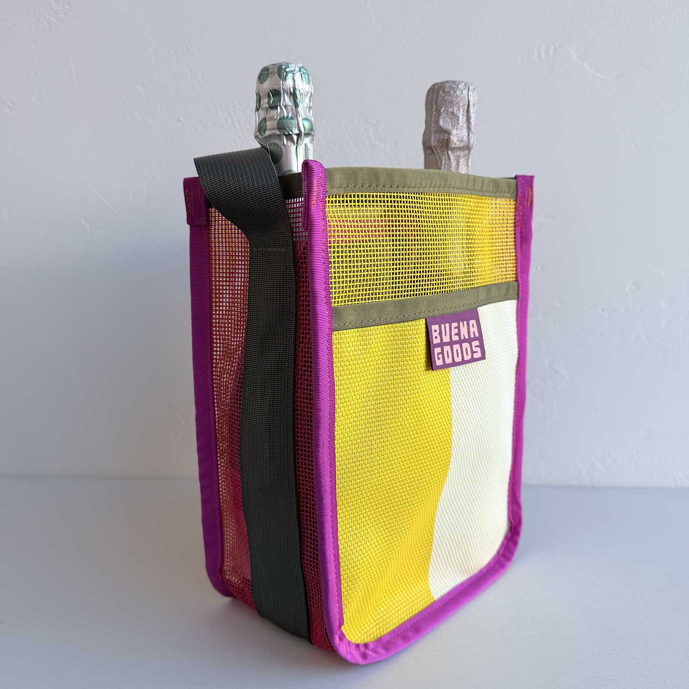 The Two-Bottle-Bag | Cava on the Playa is a colorful tote labeled “BUENA GOODS,” with purple trim and yellow, white, and burgundy panels. It holds two bottles—one in green foil, one in silver—for a stylish wine carry.