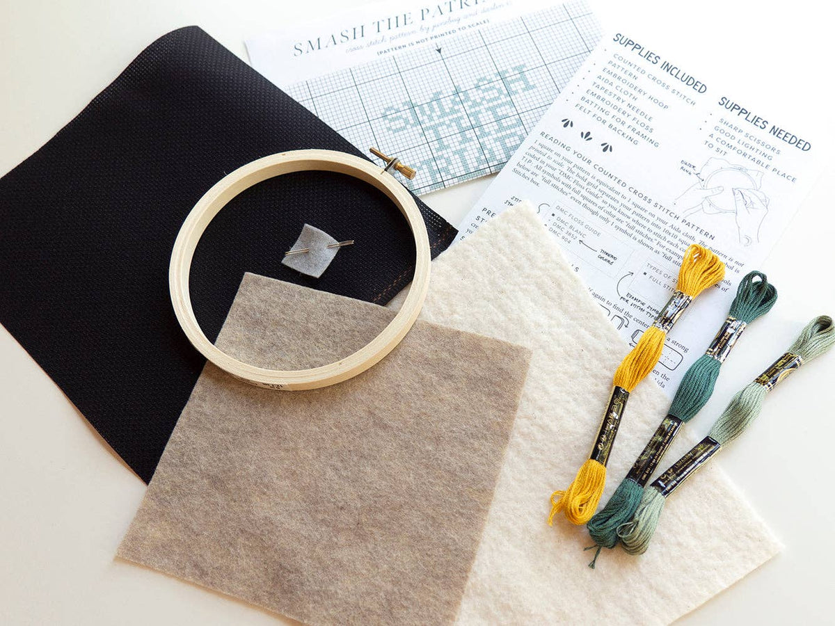 Smash the Patriarchy Cross Stitch Kit includes fabric squares, hoop, pattern instructions, and four skeins of yellow, green, and blue floss—all arranged on a white surface. Perfect for beginners and creative cross stitch projects.