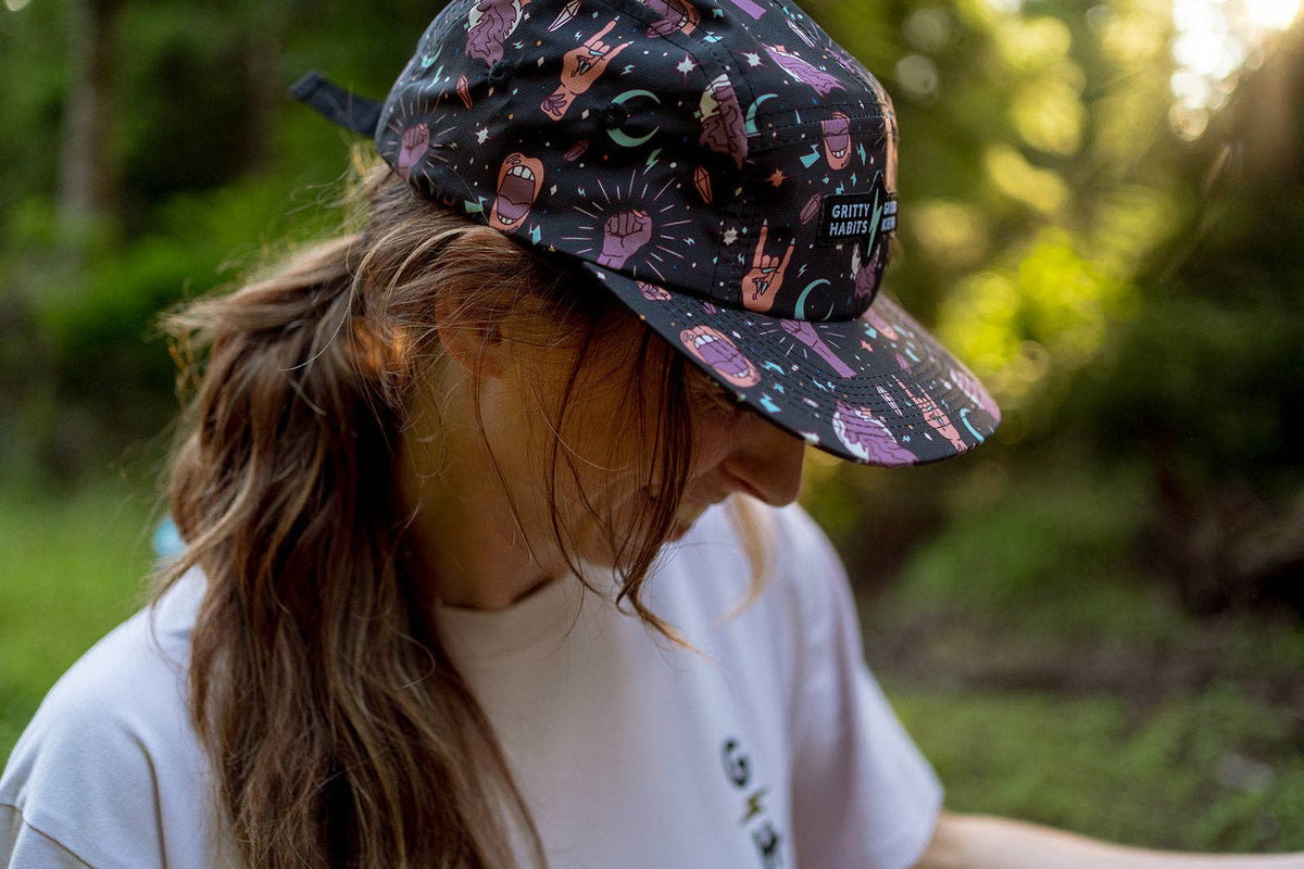 A person with long brown hair in a ponytail looks down while wearing the Gritty Habits x Ground Keeper: Howl Collection 5-Panel Hat, featuring cartoon designs, and a white shirt in a sunlit forest with lush greenery.