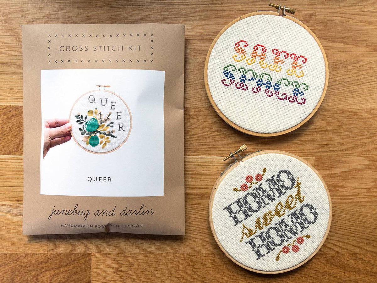A beginner Homo Sweet Homo (Foyer) Cross Stitch Kit sits on a wooden surface beside two finished hoops reading SAFE SPACE in rainbow and HOME SWEET HOME in yellow and gray—ideal for Pride Month gifts or embroidery fans.
