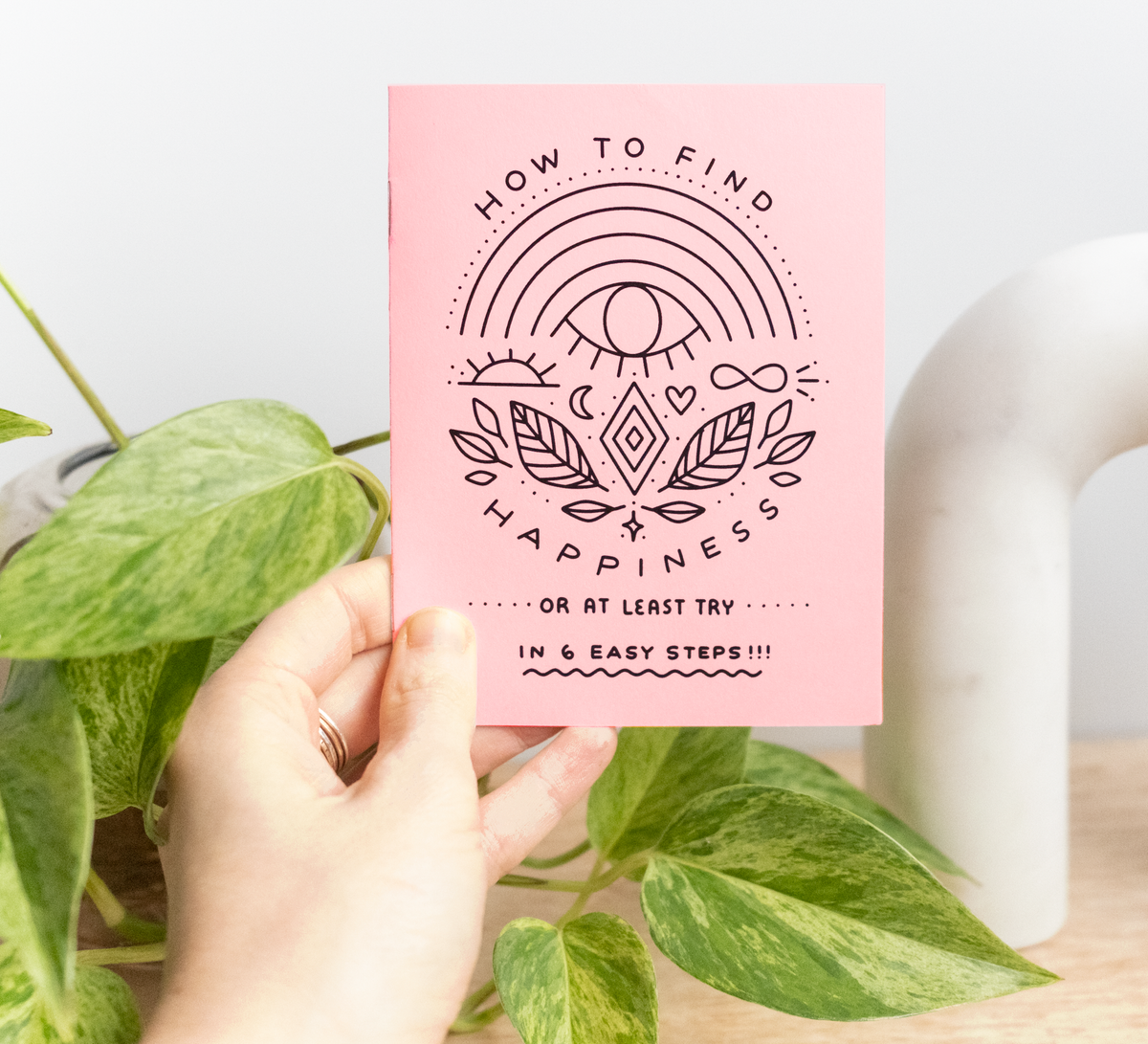 A hand holds the Little Happiness Zine, titled "How to find happiness (or at least try) in 6 easy steps," featuring black line art illustrations, with green leafy plants and a white object in the background.