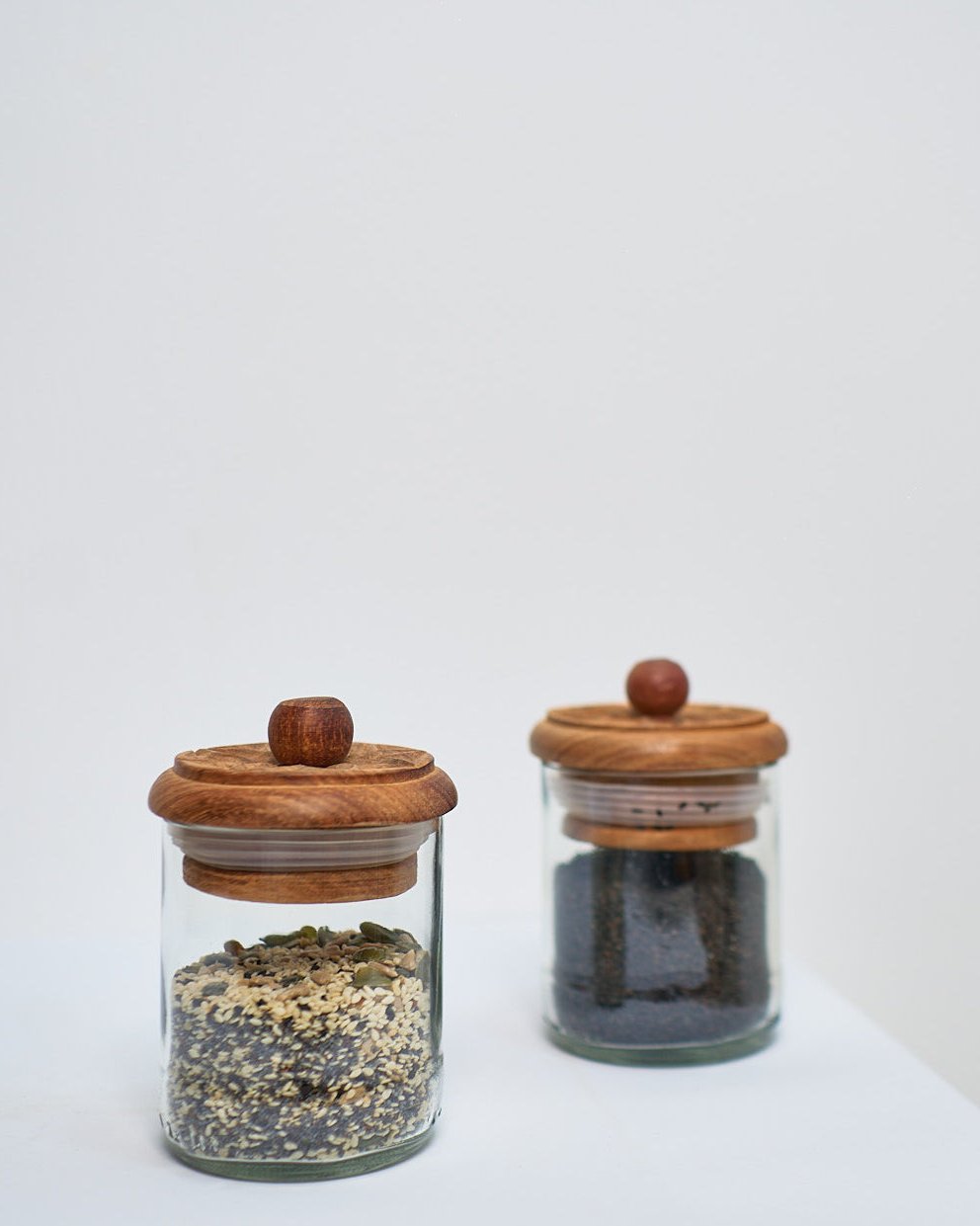 Two Karafuu Upcycled Glass Jars | Medium, each with a carved wooden lid, sit on a white surface—one filled with seeds, the other with dark Zanzibar cloves. Their minimalist design contrasts beautifully against the plain background.