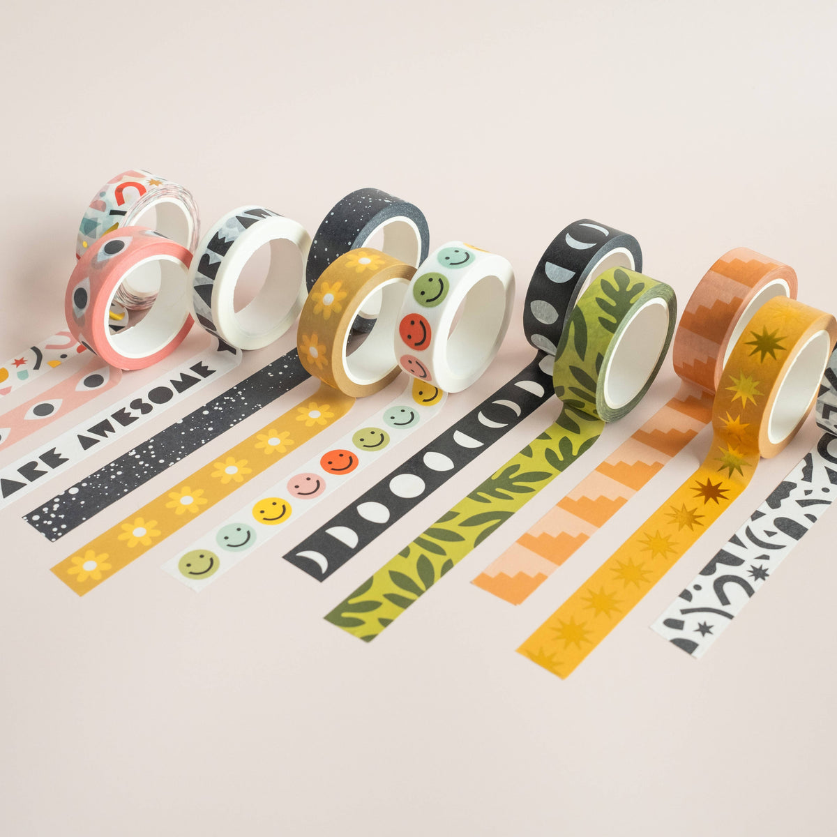 Eight rolls of Colorblock Steps Washi Tape, each featuring a unique pattern—such as smiley faces, stars, leaves, flowers, and geometric designs—are shown unrolled on a light pink surface and come in eco-friendly packaging.