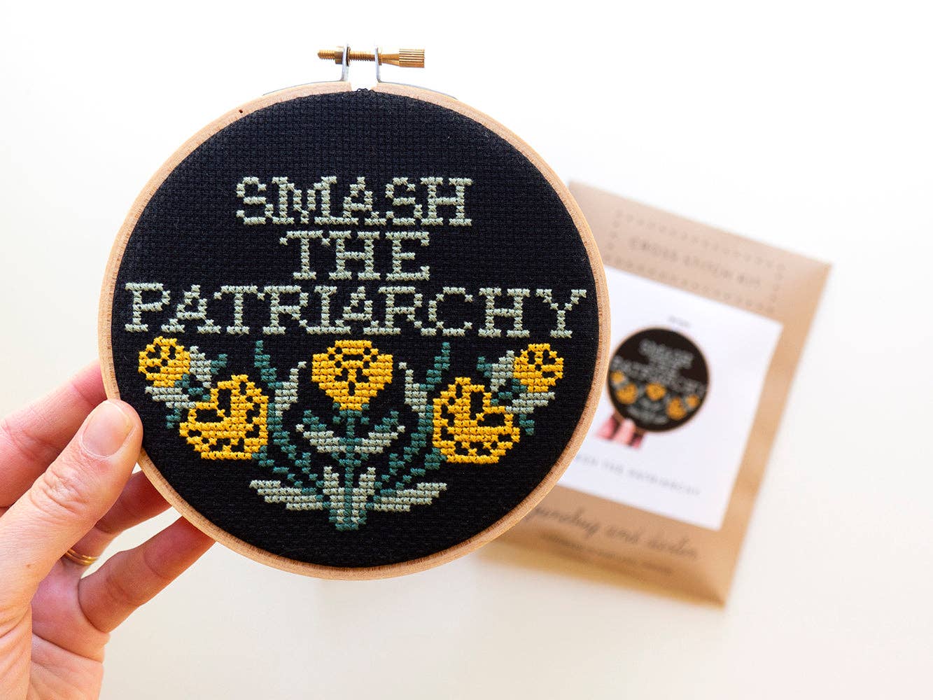 A hand holds the beginner-friendly Smash the Patriarchy Cross Stitch Kit, featuring an embroidery hoop with black fabric, yellow flowers, and “SMASH THE PATRIARCHY” stitched in white; a matching kit is visible in the background.