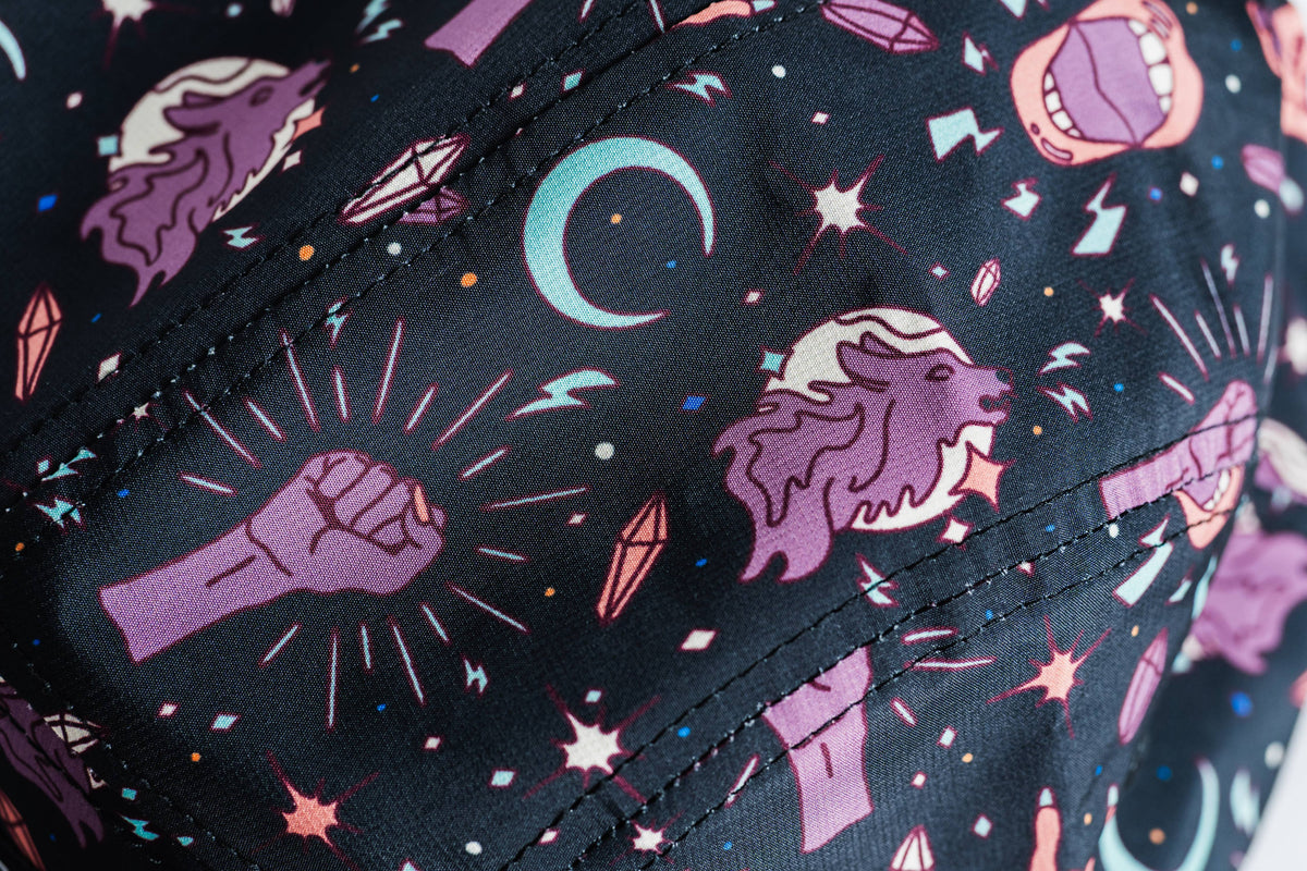 A close-up of fabric from the Gritty Habits x Ground Keeper: Howl Collection 5-Panel Hat, showing purple fists, roaring lion heads, crescent moons, pink crystals, stars, and lightning bolts on a dark background.