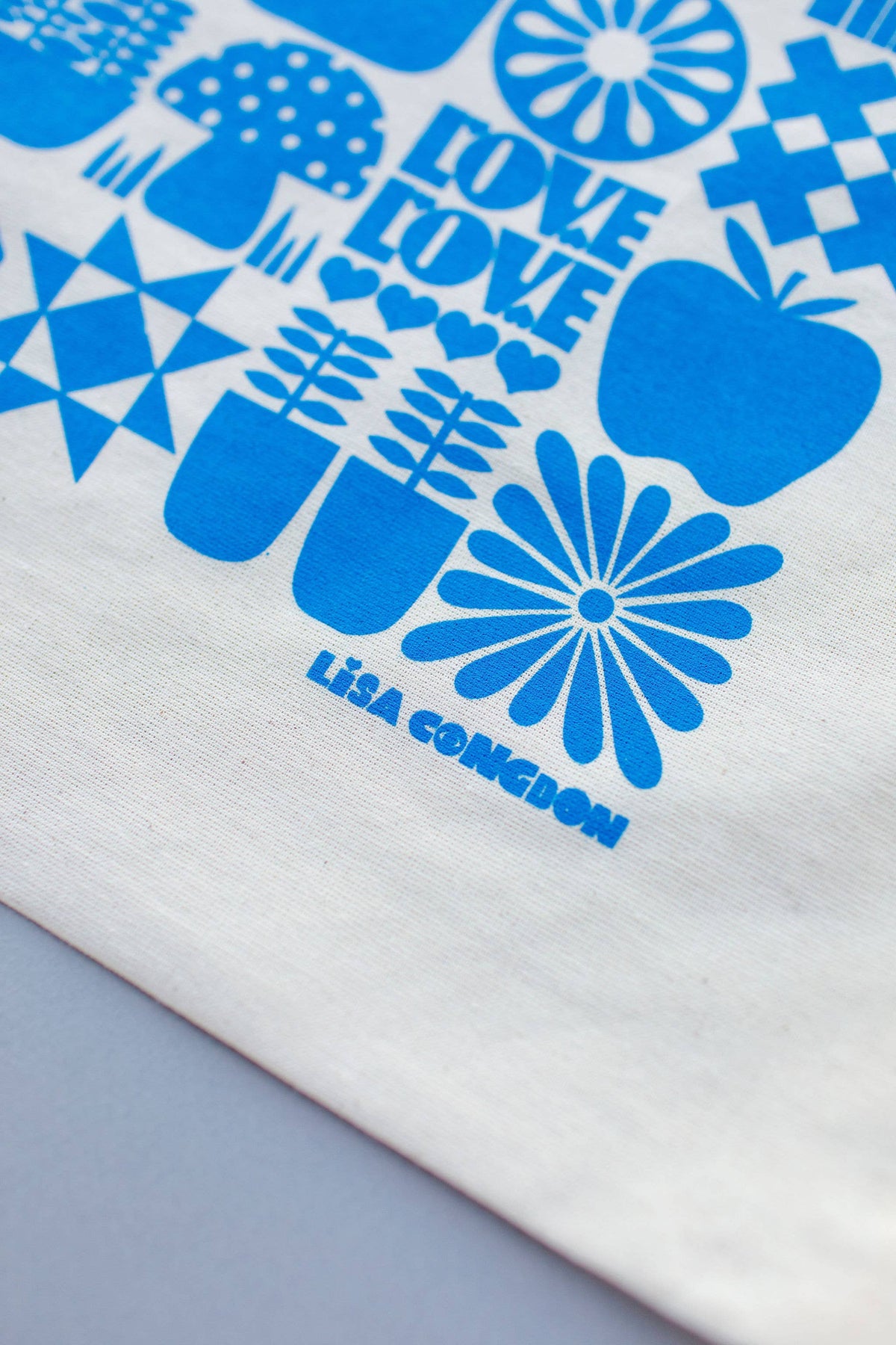 A close-up of the Large Cotton Drawstring Pouch, a white reusable gift bag with bright blue flowers, apples, stars, and "LOVE" printed twice; Lisa Congdon’s name appears at the bottom.