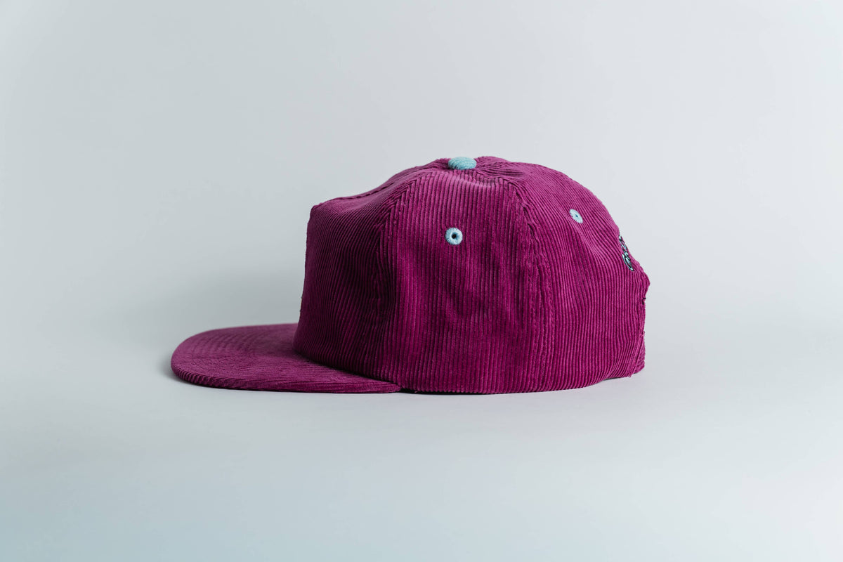 The Shred Til She's Dead Corduroy 5-Panel Hat is magenta with a flat brim, blue ventilation eyelets, a light blue top button, and a moisture-wicking sweatband. Shown from the side against a plain white background.