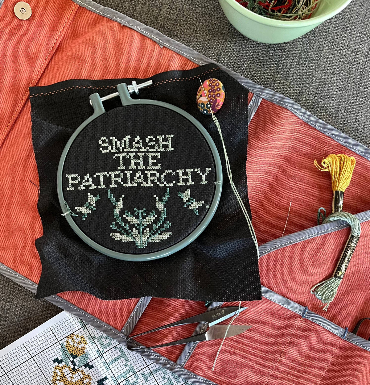 The Smash the Patriarchy Cross Stitch Kit includes a hoop project with "SMASH THE PATRIARCHY" on black fabric, decorative leaves, embroidery floss, scissors, and a pattern chart—perfect for beginners.