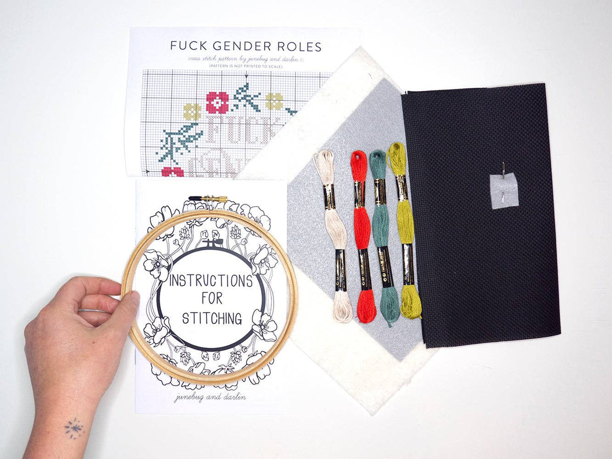 A hand holds the Fuck Gender Roles Cross Stitch Kit with a paper titled “INSTRUCTIONS FOR STITCHING.” Nearby are embroidery threads, fabric, a needle, and a beginner-friendly floral pattern labeled "FUCK GENDER ROLES.