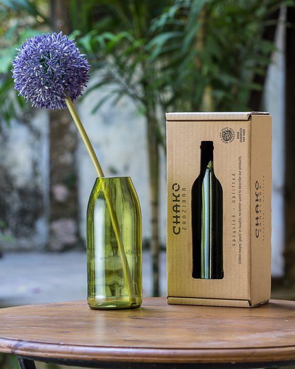 The Kikuba Upcycled Vase, handcrafted in green glass in Zanzibar, holds a single purple allium beside a brown “CHAKO Zanzibar” box on a wooden outdoor table with greenery—a stylish and sustainable home decor piece.