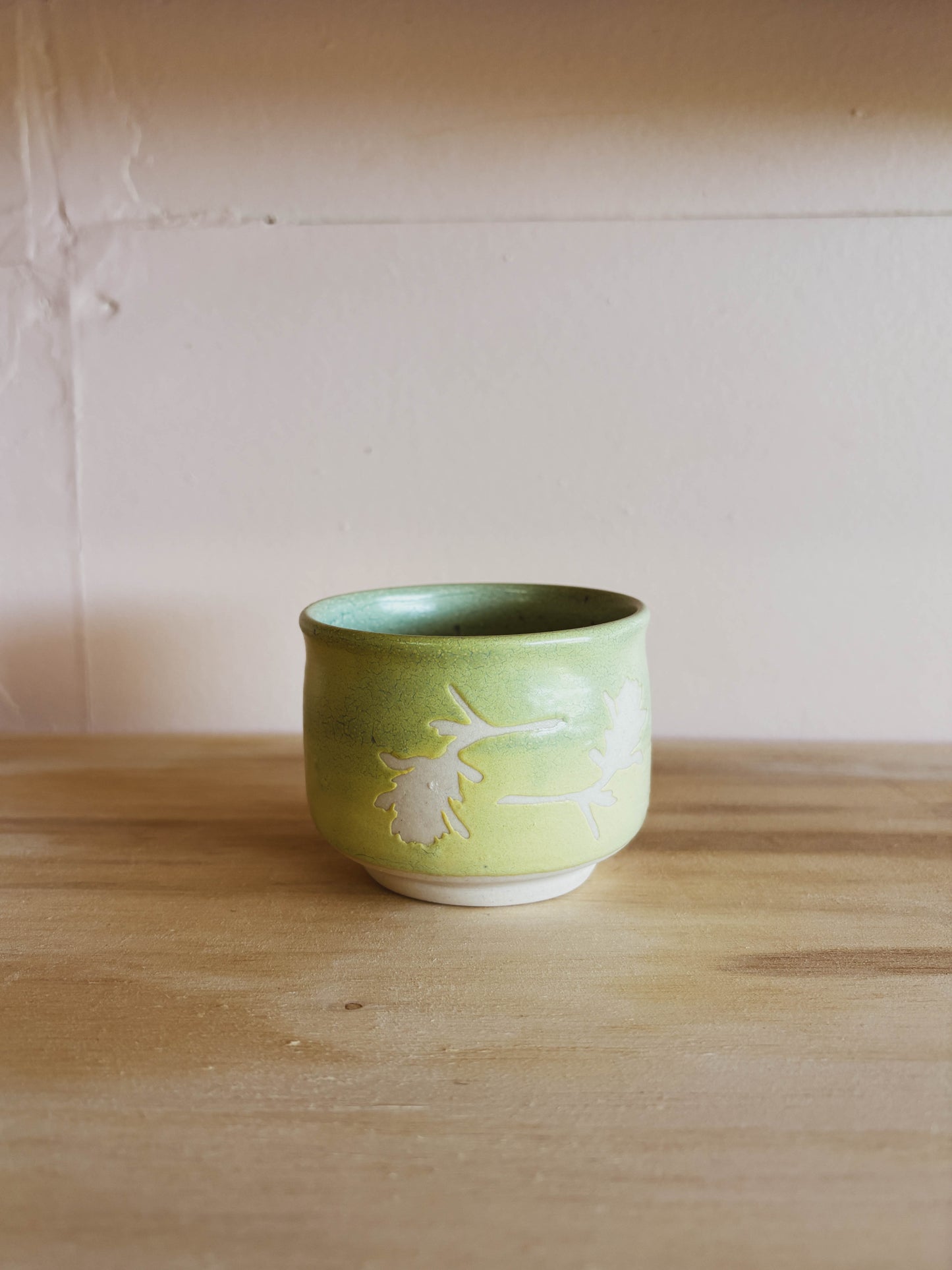 The Jade Paintbrush #1 Mini Tea Cup, a small handcrafted ceramic cup featuring a white leaf design, rests on a light wooden surface against a pale beige wall—an elegant choice for enjoying mini servings of tea.