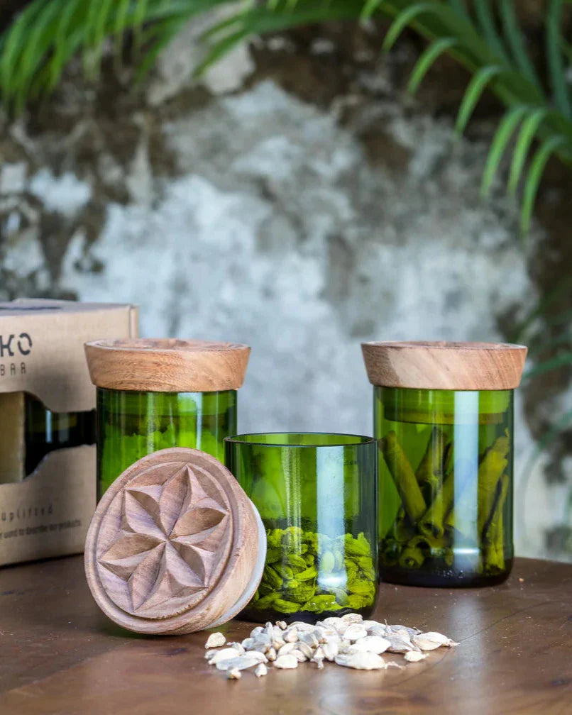 The Hiliki Upcycled Glass Jars | Large Set features three glass jars with carved wooden lids—two holding dried herbs or spices. One lid showcases its pattern beside scattered seeds and pods, while a leafy plant sits before a stone wall.