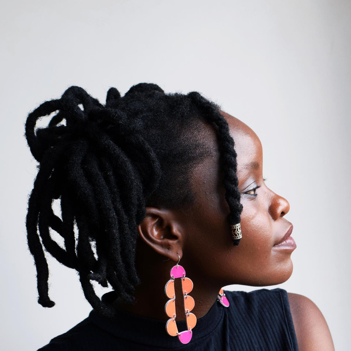 A woman with black styled locs wears vibrant Sisanda Paper Pulp Earrings from Quazi Design and a sleeveless black top, shown in profile against a plain white background, looking to the right.