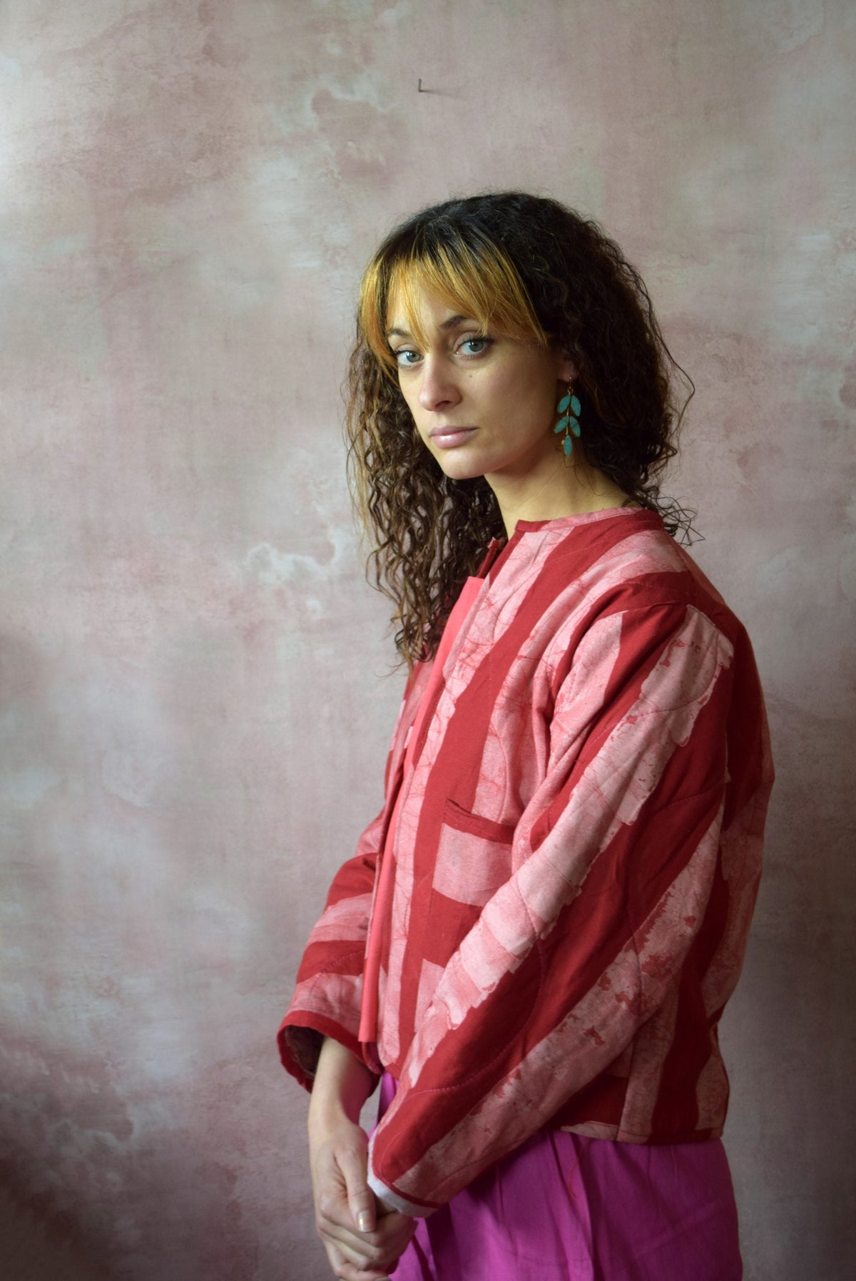 A woman with curly hair wears Posy Paper Pulp Earrings and a red and pink patterned top with magenta pants, standing against a pink textured background, looking thoughtfully at the camera.