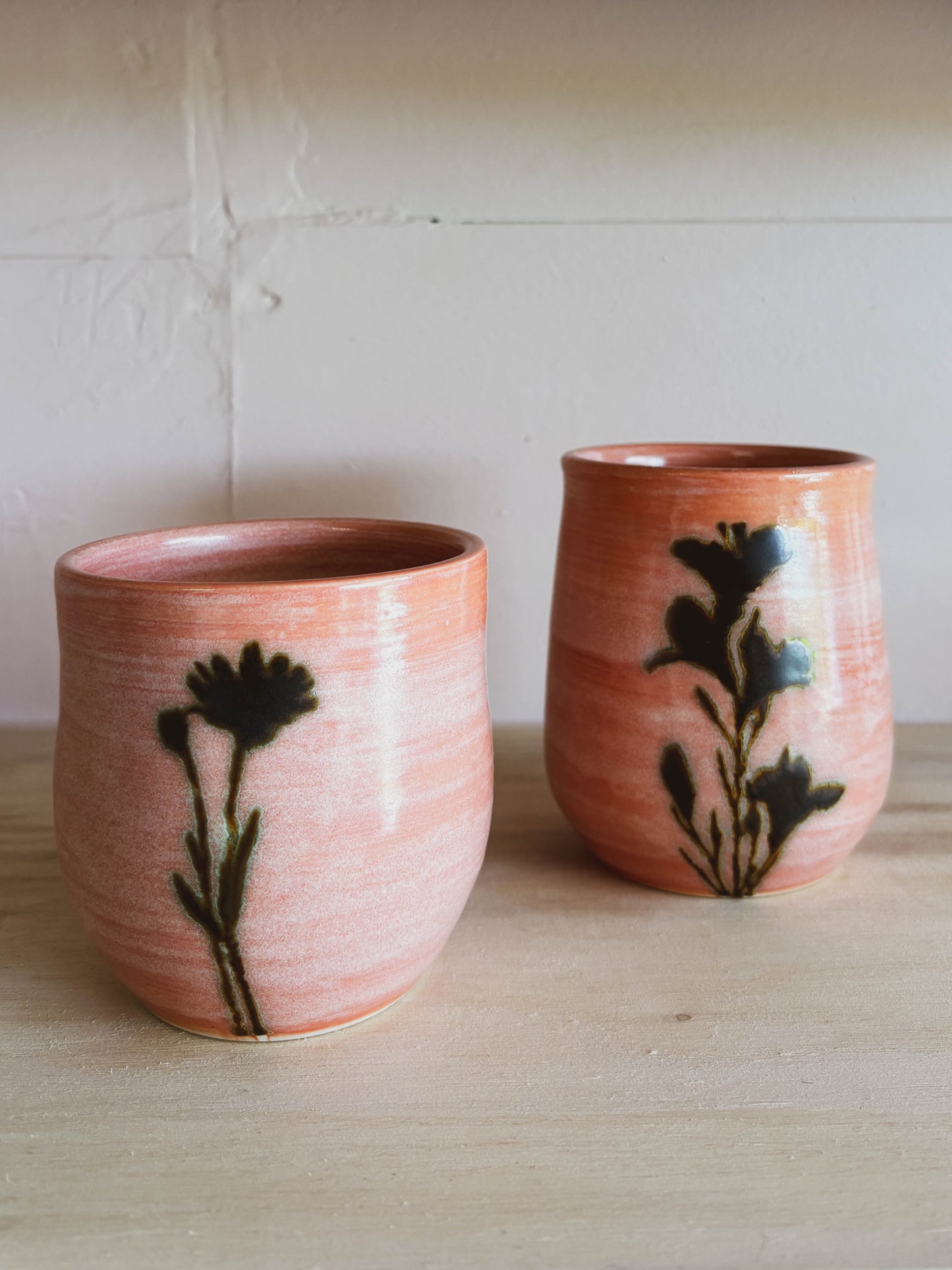 The Rose Quartz Arnica #2 Medium Tea Cup features a coral hue with black floral designs. Handcrafted from ceramic, this unique cup adds charm and elegance to any table setting.