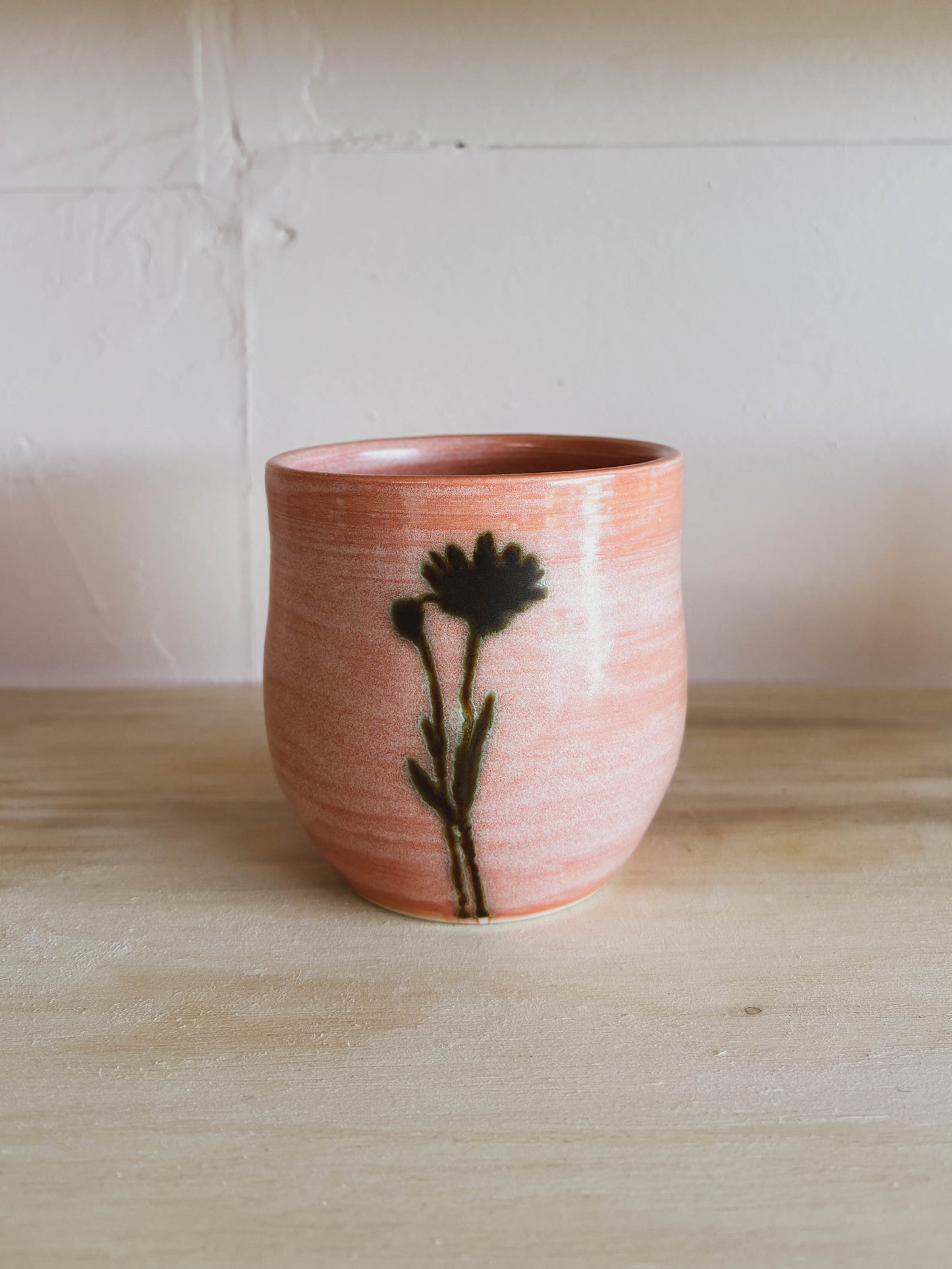 The Rose Quartz Arnica #2 Medium Tea Cup features a handcrafted ceramic body with a soft pink glaze and dark floral silhouette, displayed on light wood against an off-white background—making it a truly unique tea cup.