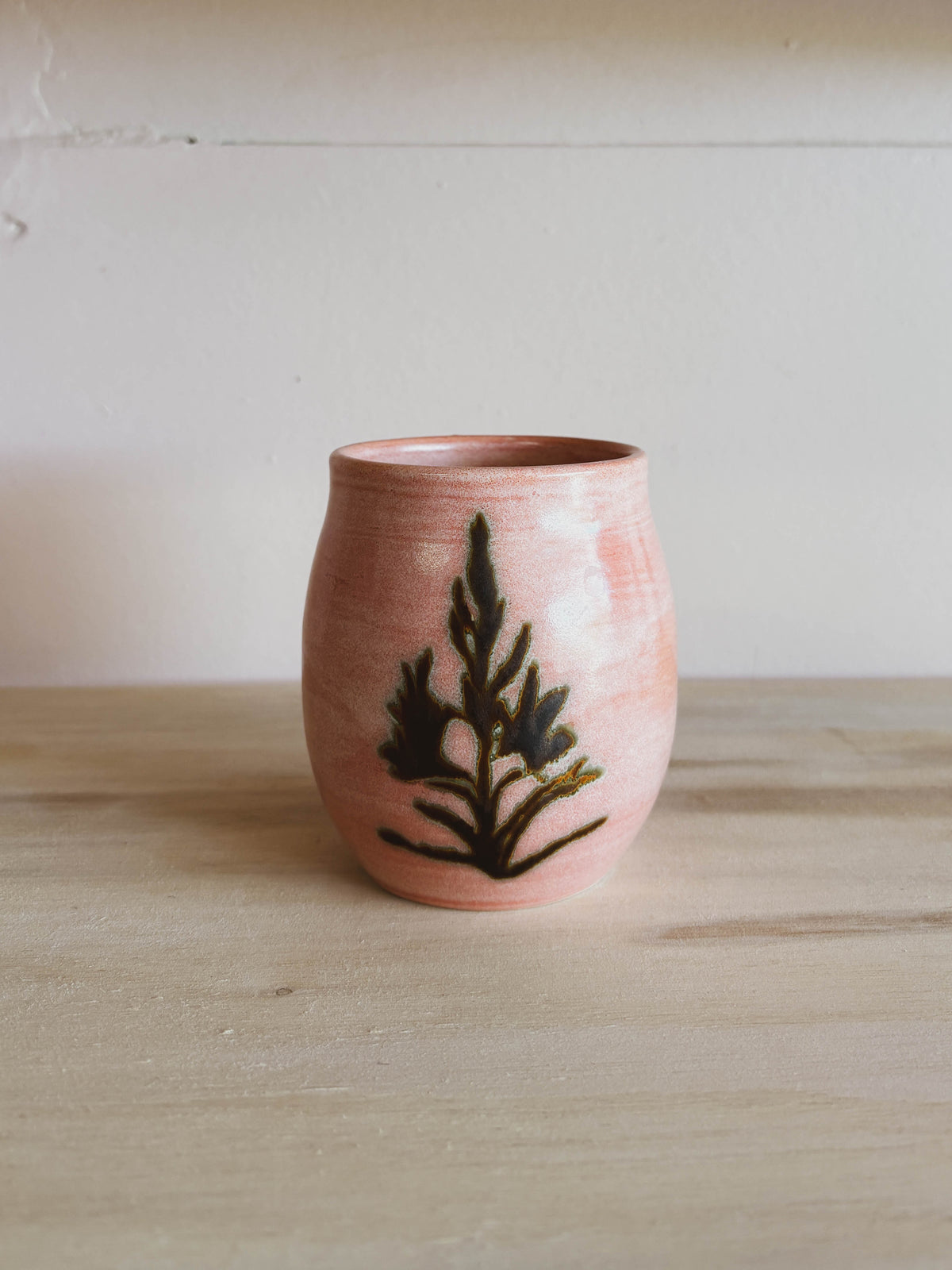 The Rose Quartz Lily #2 Small Tea Cup, a pink handcrafted ceramic cup with a rounded shape and dark green leafy design, rests on a light wooden surface against an off-white background.
