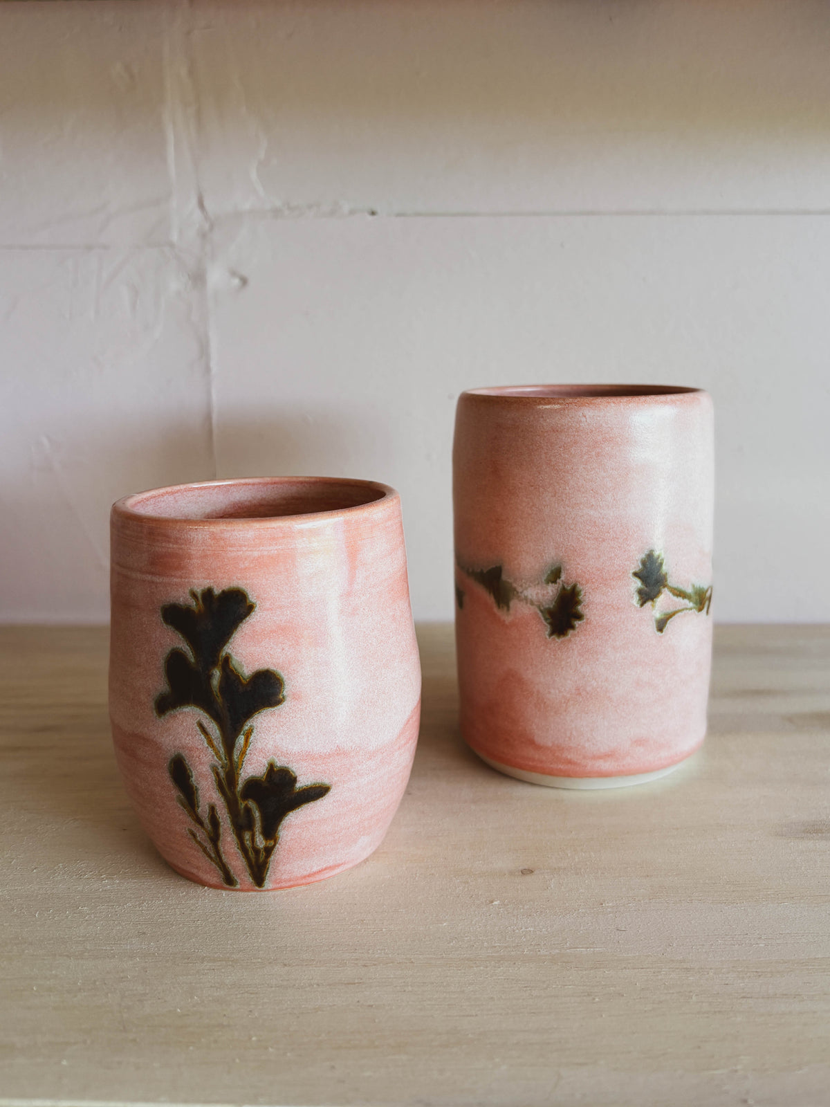 The Rose Quartz Lily #1 Small Tea Cups, handcrafted in soft pink with dark botanical designs, rest on a light wooden surface by a white wall. These elegant, women-owned pieces are dishwasher safe.