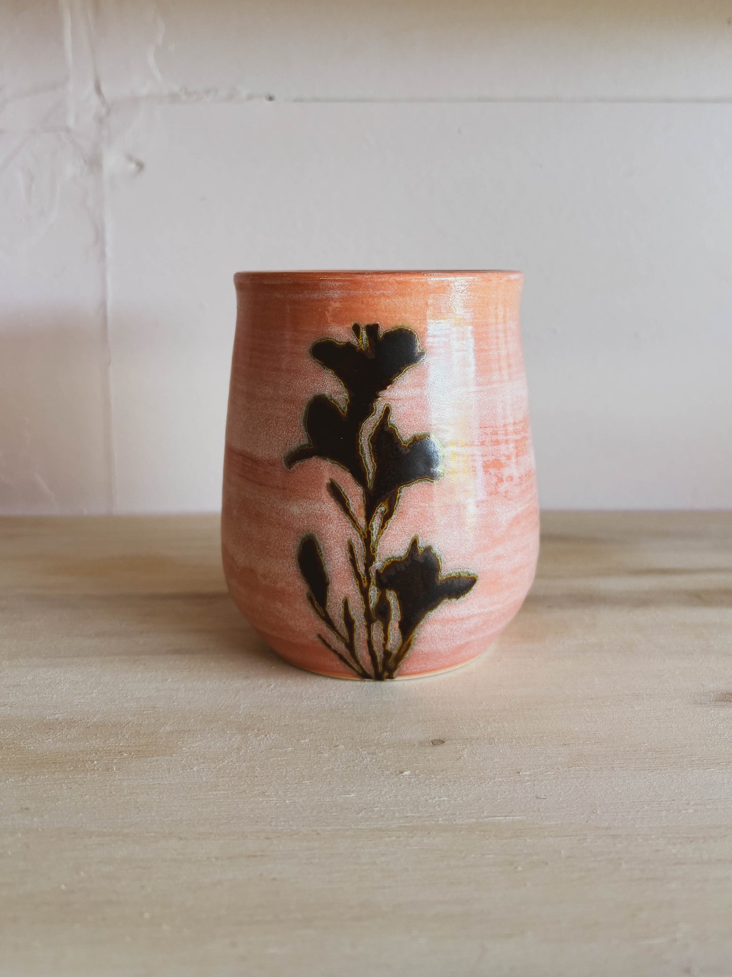 The Rose Quartz Lily #2 Medium Tea Cup features a pink marble finish and black floral silhouette, handcrafted by a women-owned brand. Uniquely designed, dishwasher safe, and shown on a light wood surface against a pale wall.