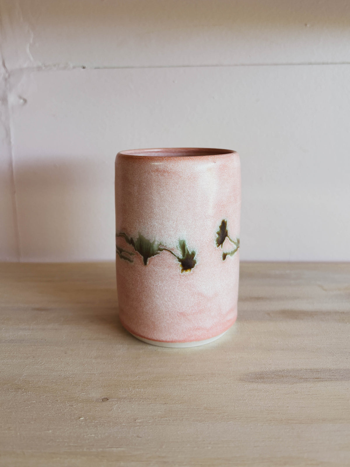 The Rose Quartz Arnica #1 Medium Tea Cup, a handcrafted ceramic piece in pink with green and brown branch-like accents, sits on light wood. This women-owned tea cup is beautiful and safe for dishwasher use.