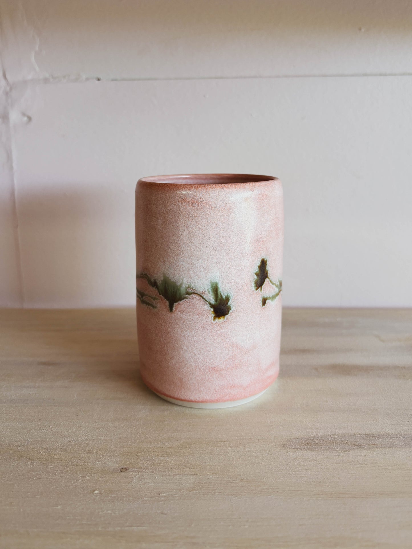 The Rose Quartz Arnica #1 Medium Tea Cup, a handcrafted ceramic piece in pink with green and brown branch-like accents, sits on light wood. This women-owned tea cup is beautiful and safe for dishwasher use.