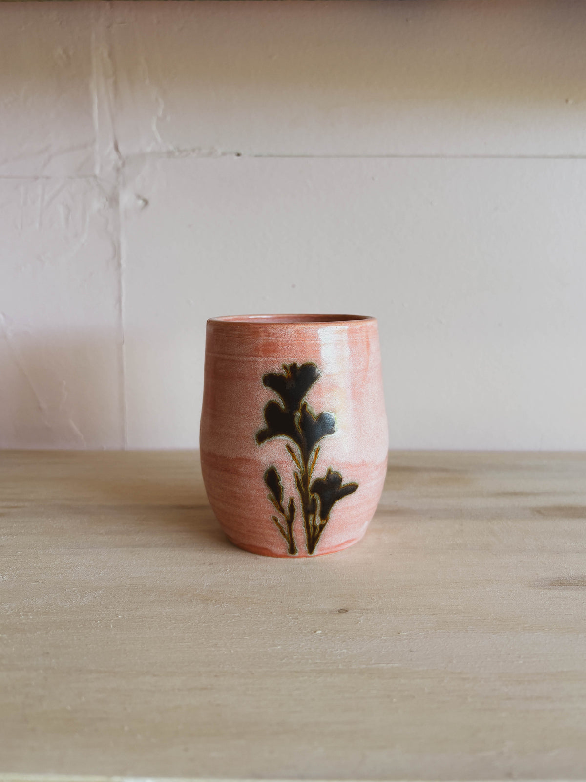 The Rose Quartz Lily #1 Small Tea Cup, a handcrafted pink ceramic cup with a black floral design, rests on a light wooden surface against a pale textured wall.