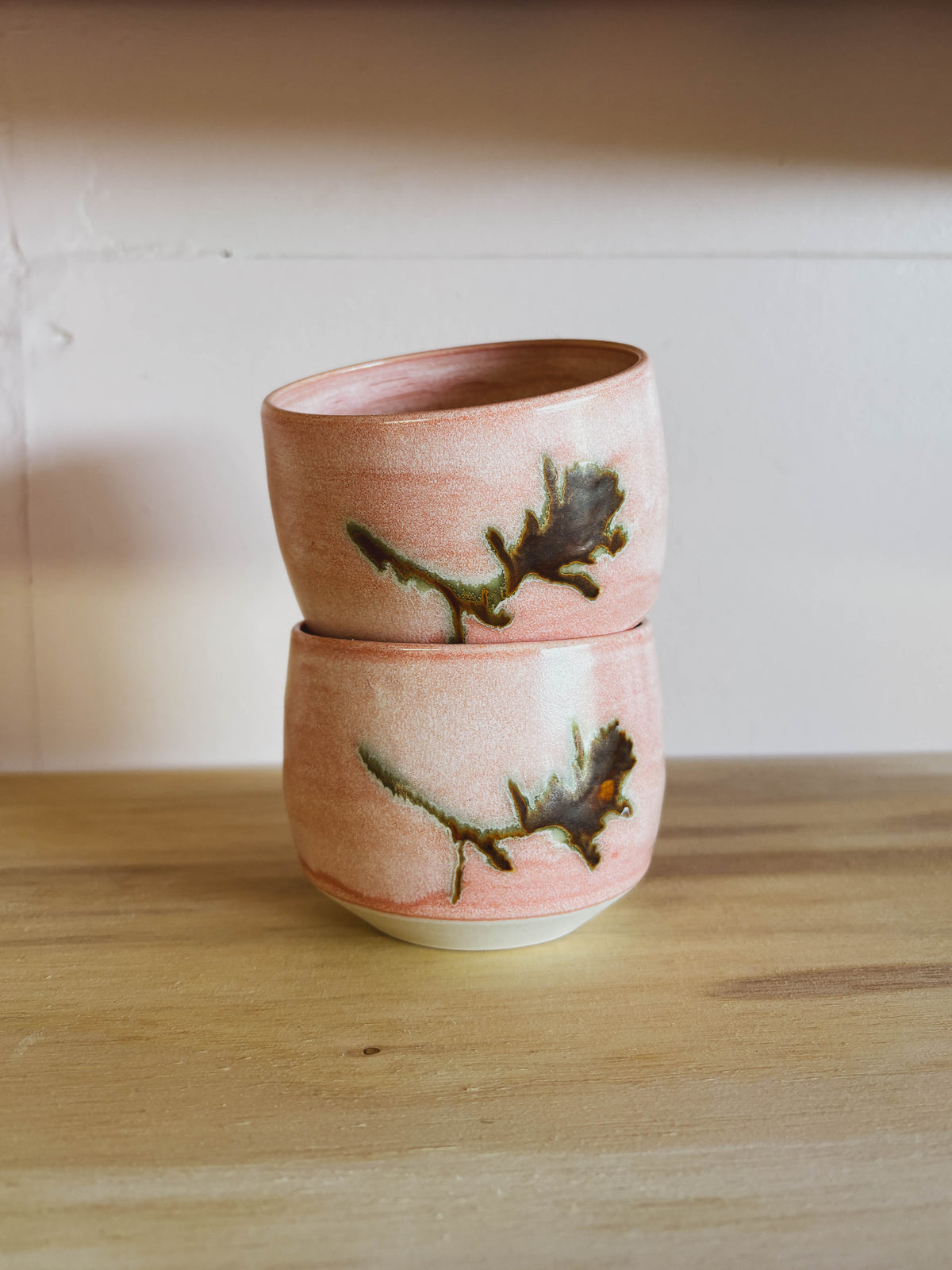 The Rose Quartz Paintbrush #1 Small Tea Cups feature a soft pink glaze and abstract greenish-brown leaf designs, stacked on light wood—unique handcrafted pieces from a women-owned studio.