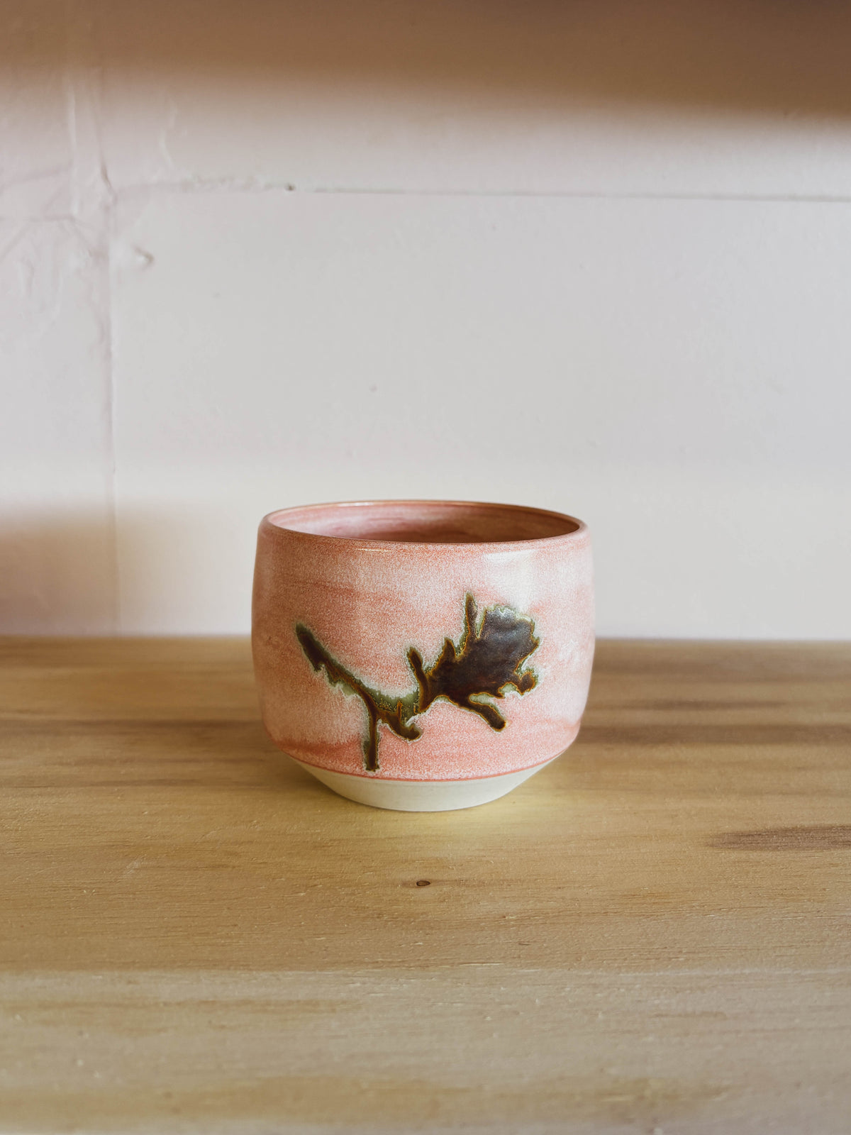 The Rose Quartz Paintbrush #2 Small Tea Cup, a handcrafted ceramic piece featuring a soft pink glaze and dark brown abstract floral design, rests on a wooden surface against a light-colored wall.