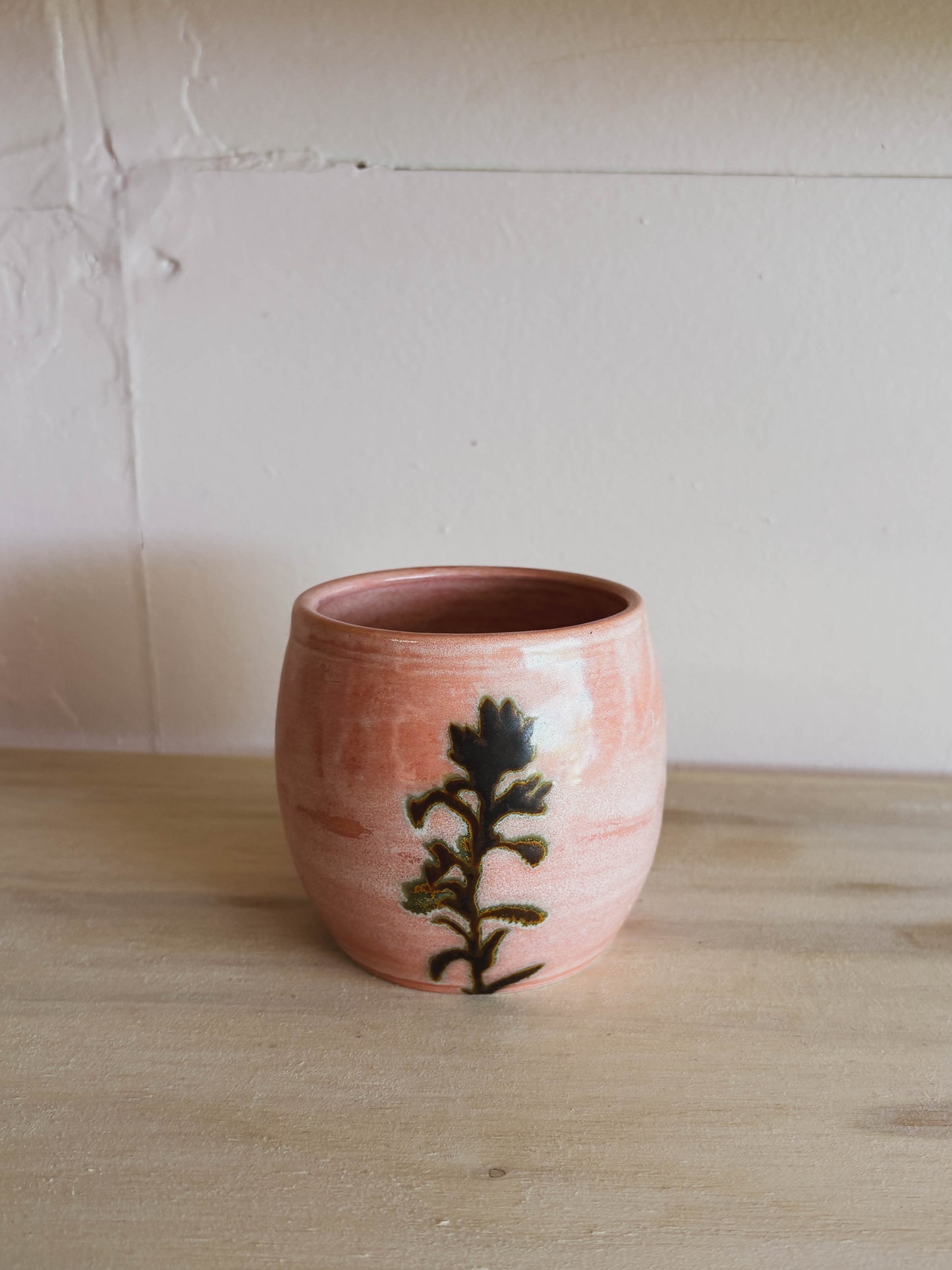 The Rose Quartz Paintbrush #4 Small Tea Cup, a handcrafted, round pink ceramic cup with a dark green leafy plant design, sits on a wooden surface against a textured light-colored wall.